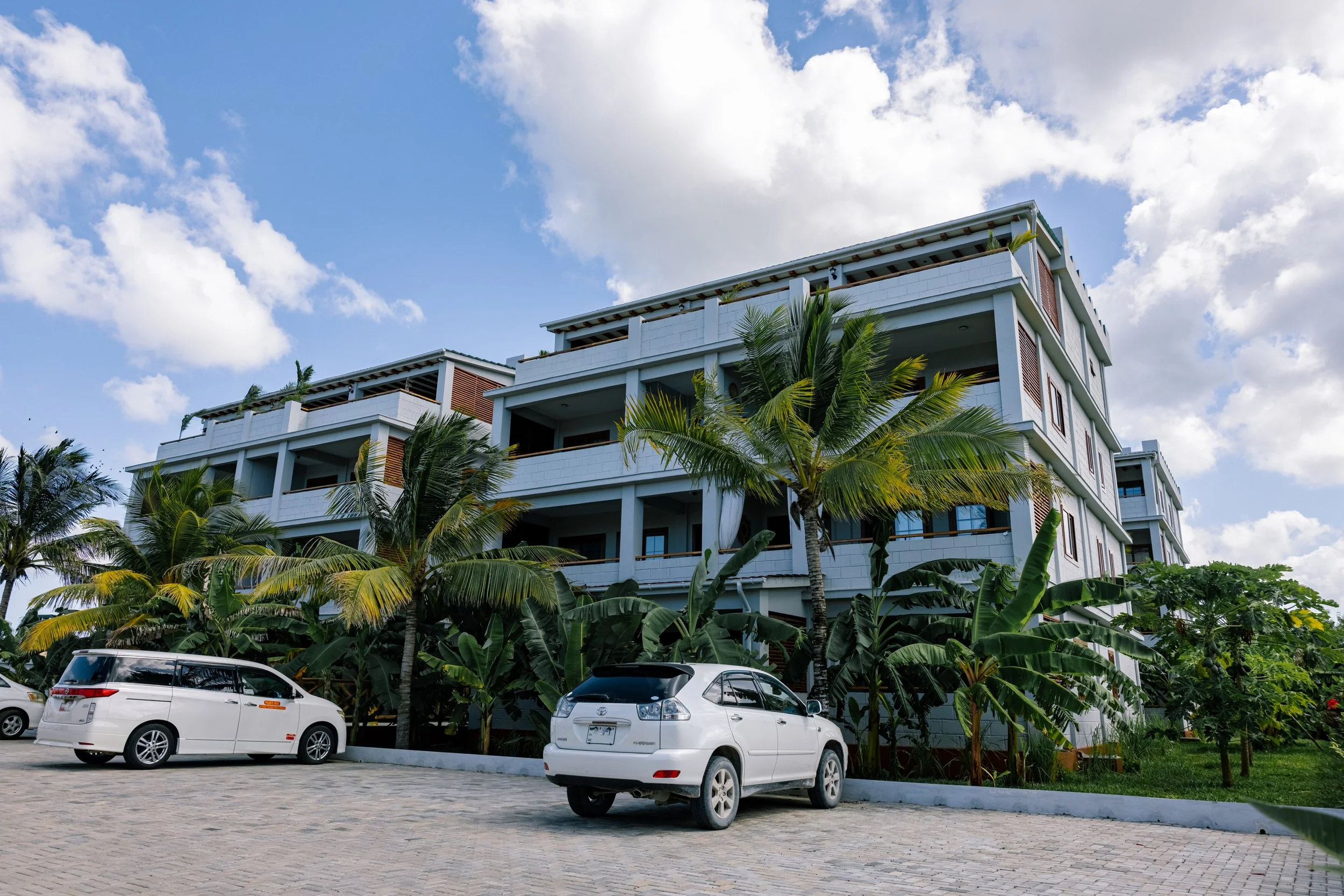 A multi-story white building with balconies, surrounded by lush palm trees, under a bright blue sky with scattered clouds. Cars are parked in front of the building on a paved lot.