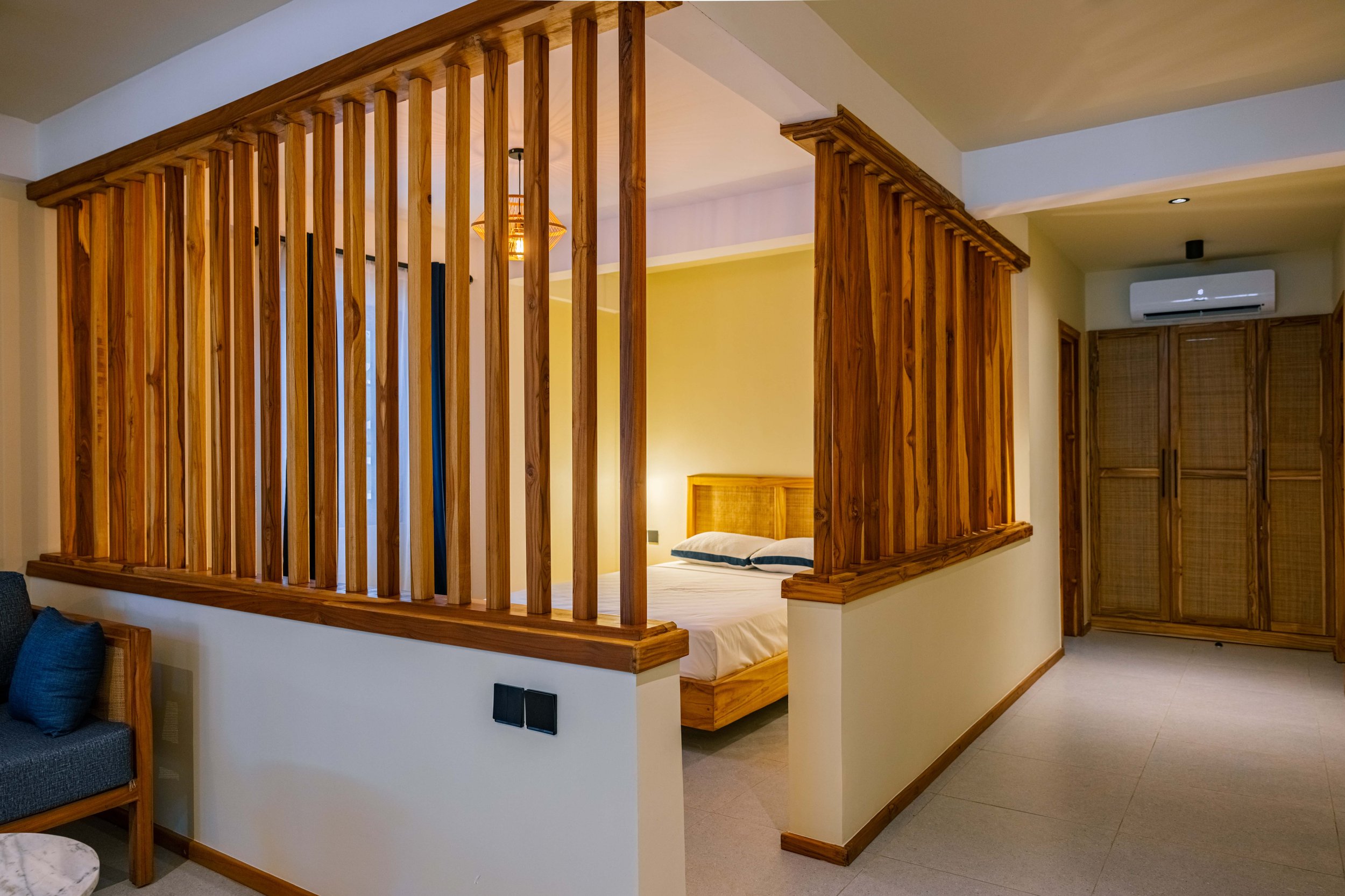 A bedroom with a wooden bed frame and wooden slatted partition separating it from the living area, with a yellow wall and a light fixture hanging from the ceiling.