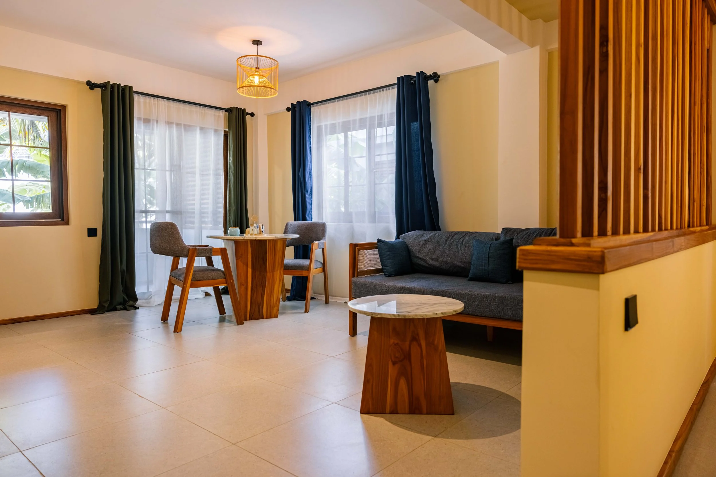 Living room with a wooden dining table, chairs, a gray sofa, large windows with curtains, a small round marble table, and a wooden room divider.