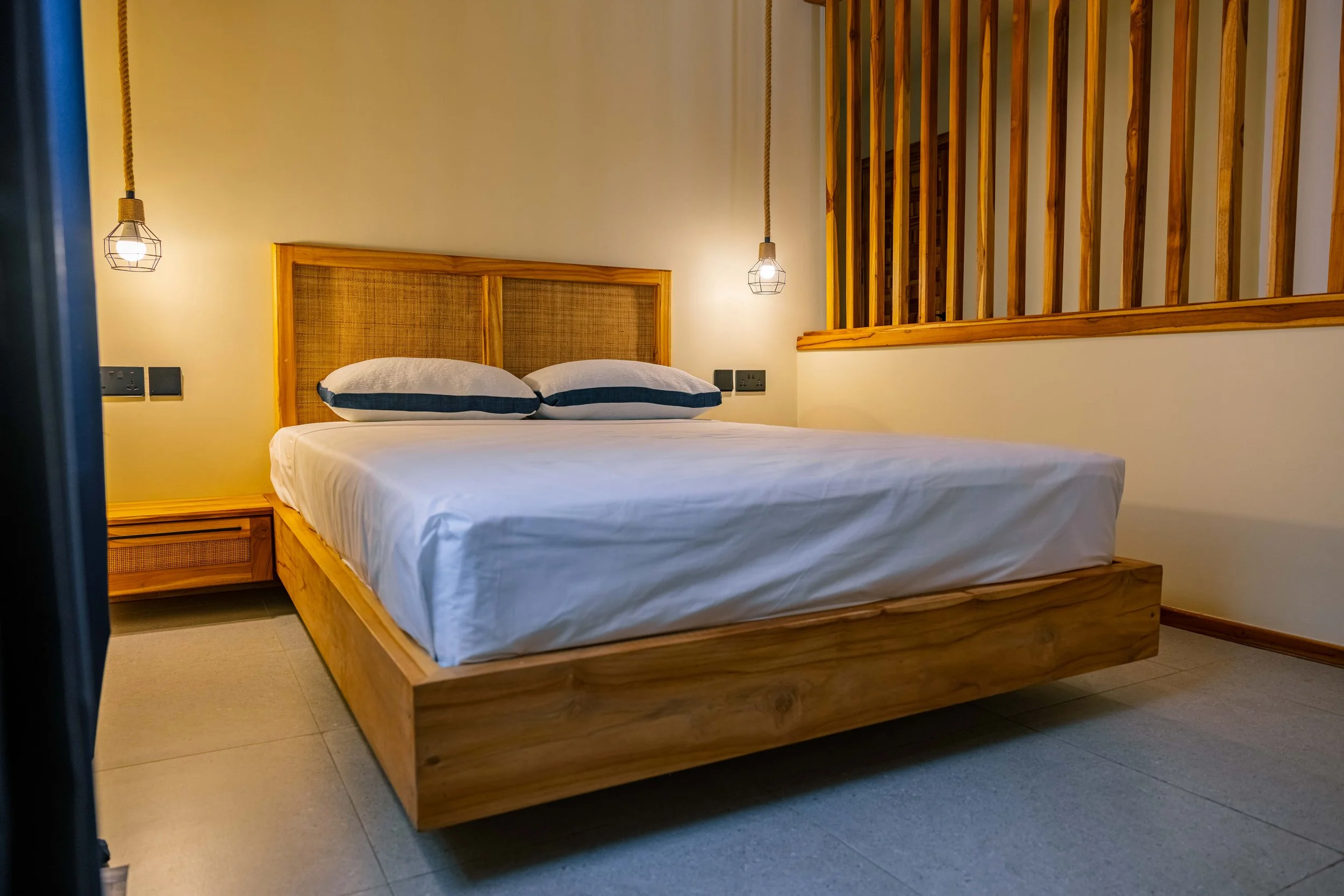 A modern bedroom with a wooden bed frame, two pillows with striped pillowcases, and wall-mounted pendant lights, with a decorative wooden partition on the right side.