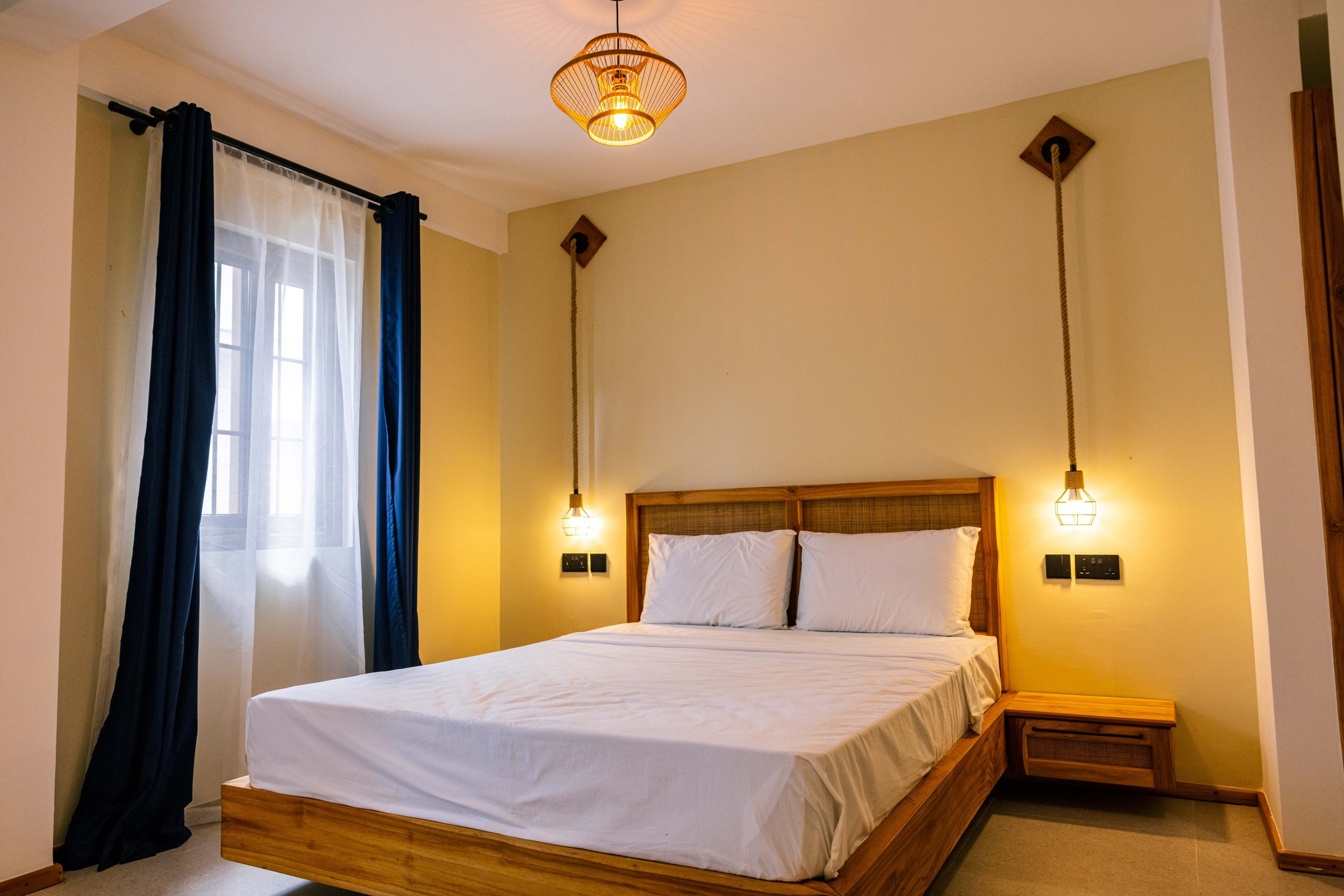 A bedroom with a wooden bed frame, white bedding, two pillows, and hanging pendant lights on both sides. There is a window with white sheer curtains and navy blue drapes, and a small wooden bedside table.