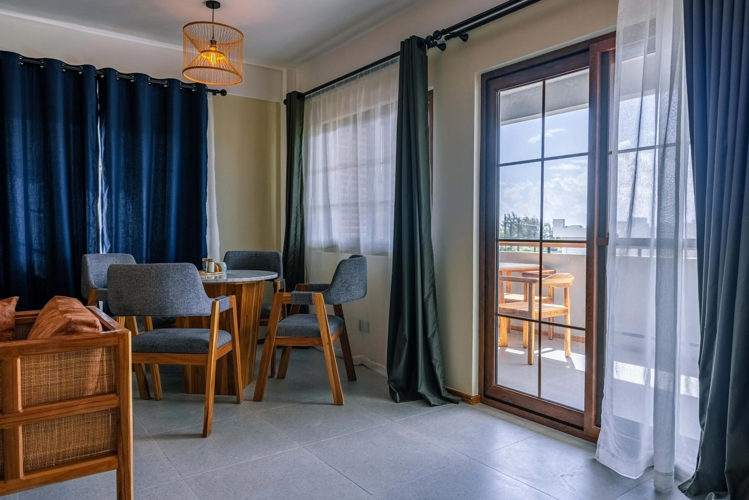 A living room with a small round dining table surrounded by gray upholstered chairs, dark blue curtains, sheer white curtains, and a sliding glass door leading to a balcony with outdoor chairs and a view of a bright sky and trees.