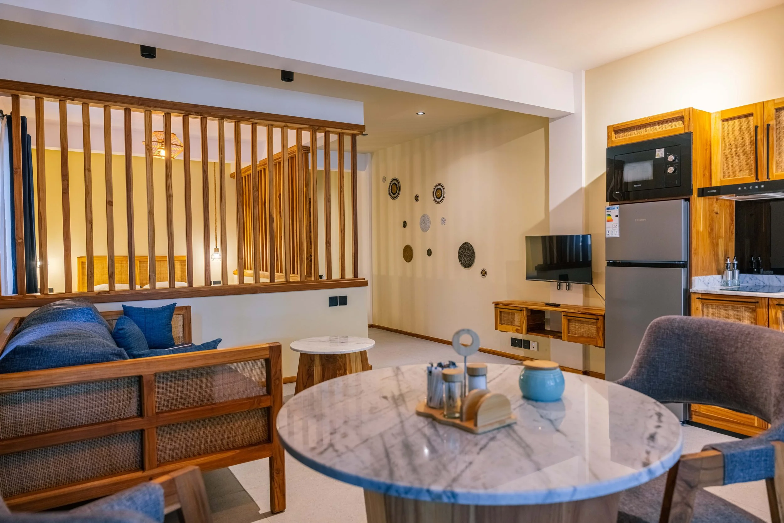 Living room with wooden furniture, a marble-top round table, a gray sofa with blue cushions, a wall TV, and kitchen appliances including a microwave and refrigerator, with wall decorations and wooden slats dividing spaces.