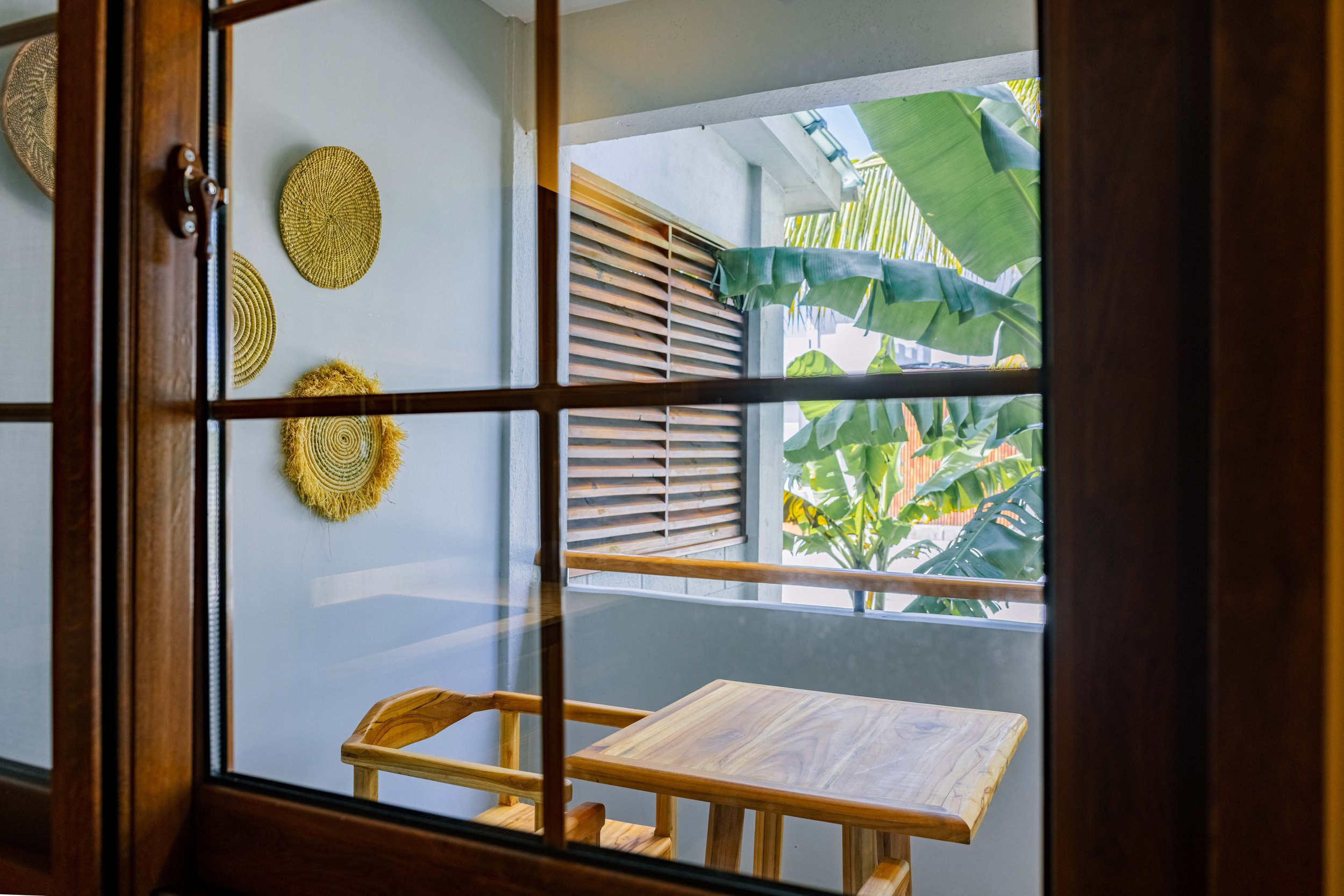 View through a window showing a small outdoor patio with a wooden table and bench, surrounded by tropical plants and a wooden privacy screen.