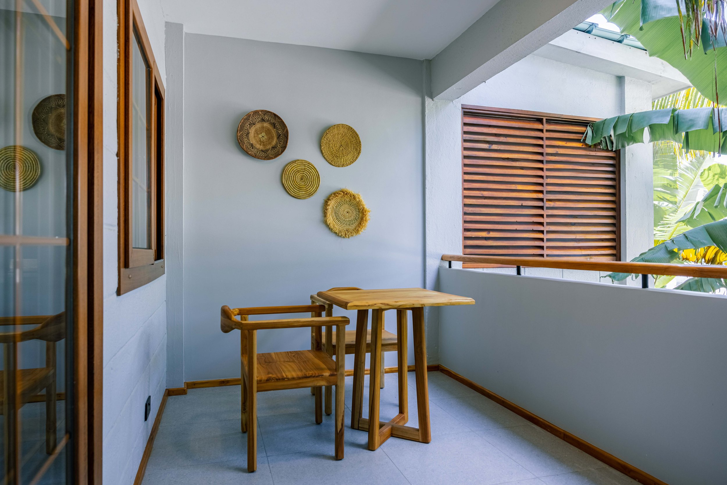 Balcony with wooden furniture, decorative woven wall art, and lush green tropical plants outside.