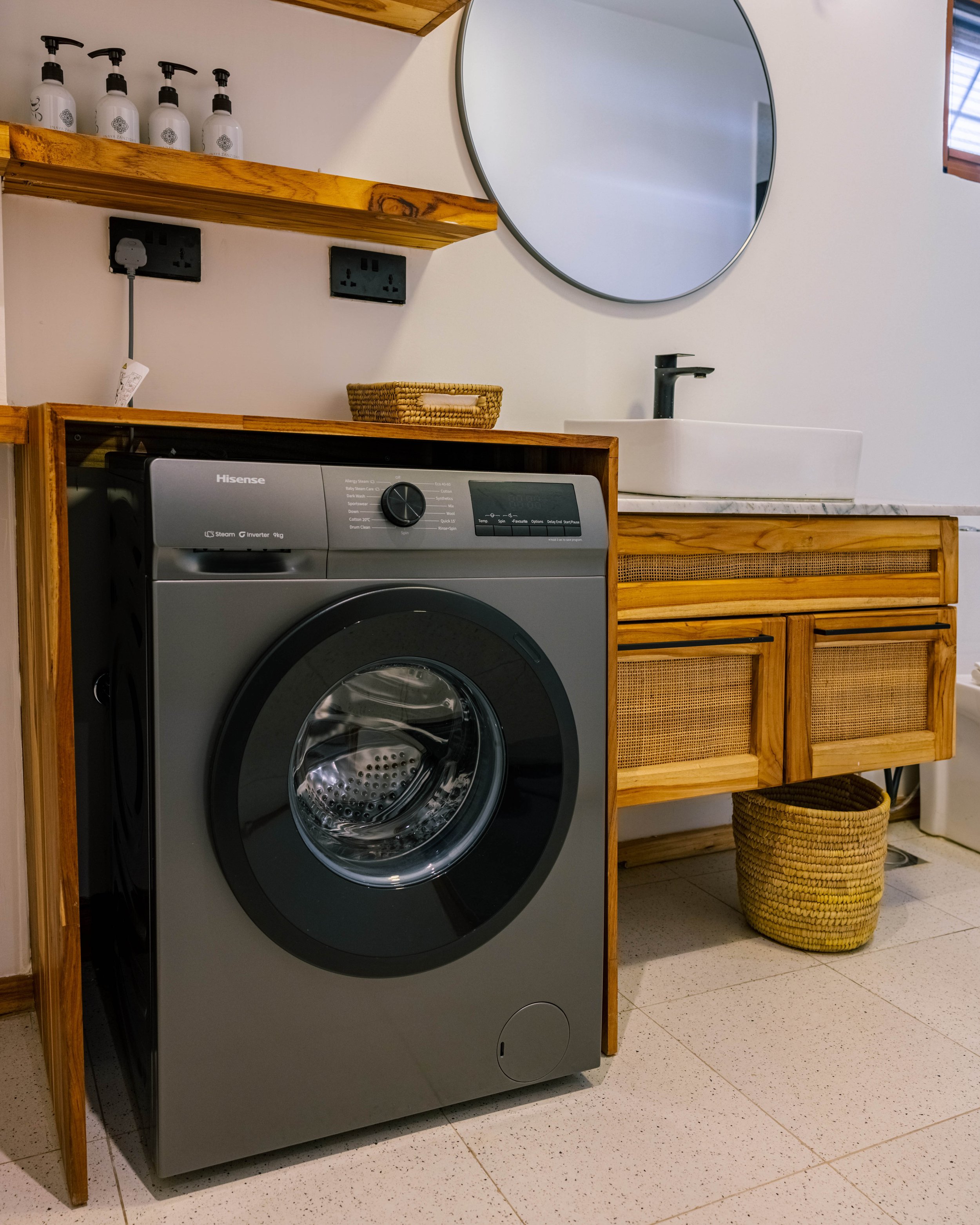 A laundry room with a front-loading clothes washer, a wooden shelf with four white bottles with pump dispensers, a nearby mirror, a small sink with a faucet, a woven basket on the floor, and a wooden cabinet with rattan panels.