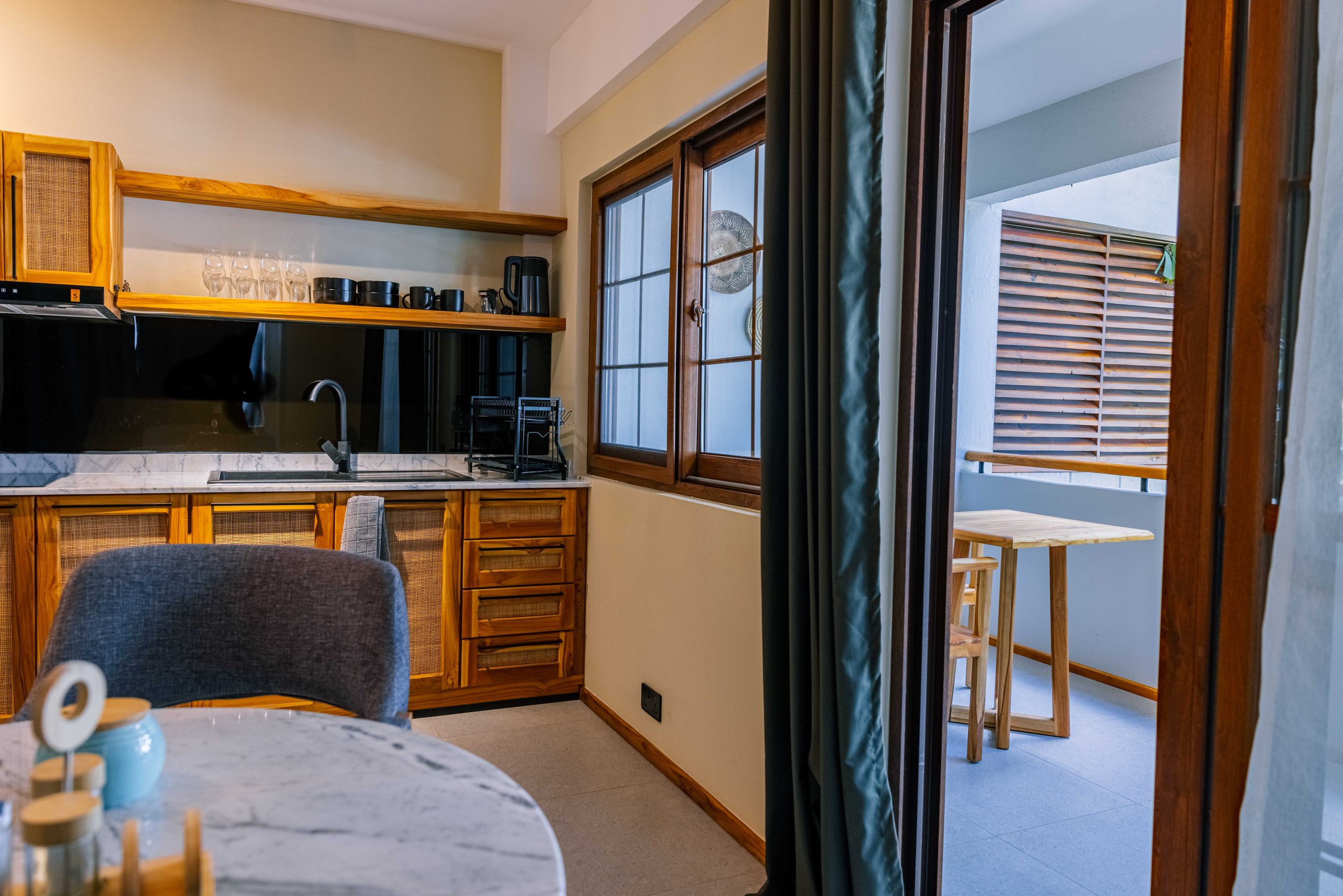 View of a cozy kitchen area with wooden cabinets, a marble countertop, and open shelves holding glassware and black cups. A window with wooden frames and partially drawn green curtains overlooks an outdoor space with wooden slats. Some chairs and a d