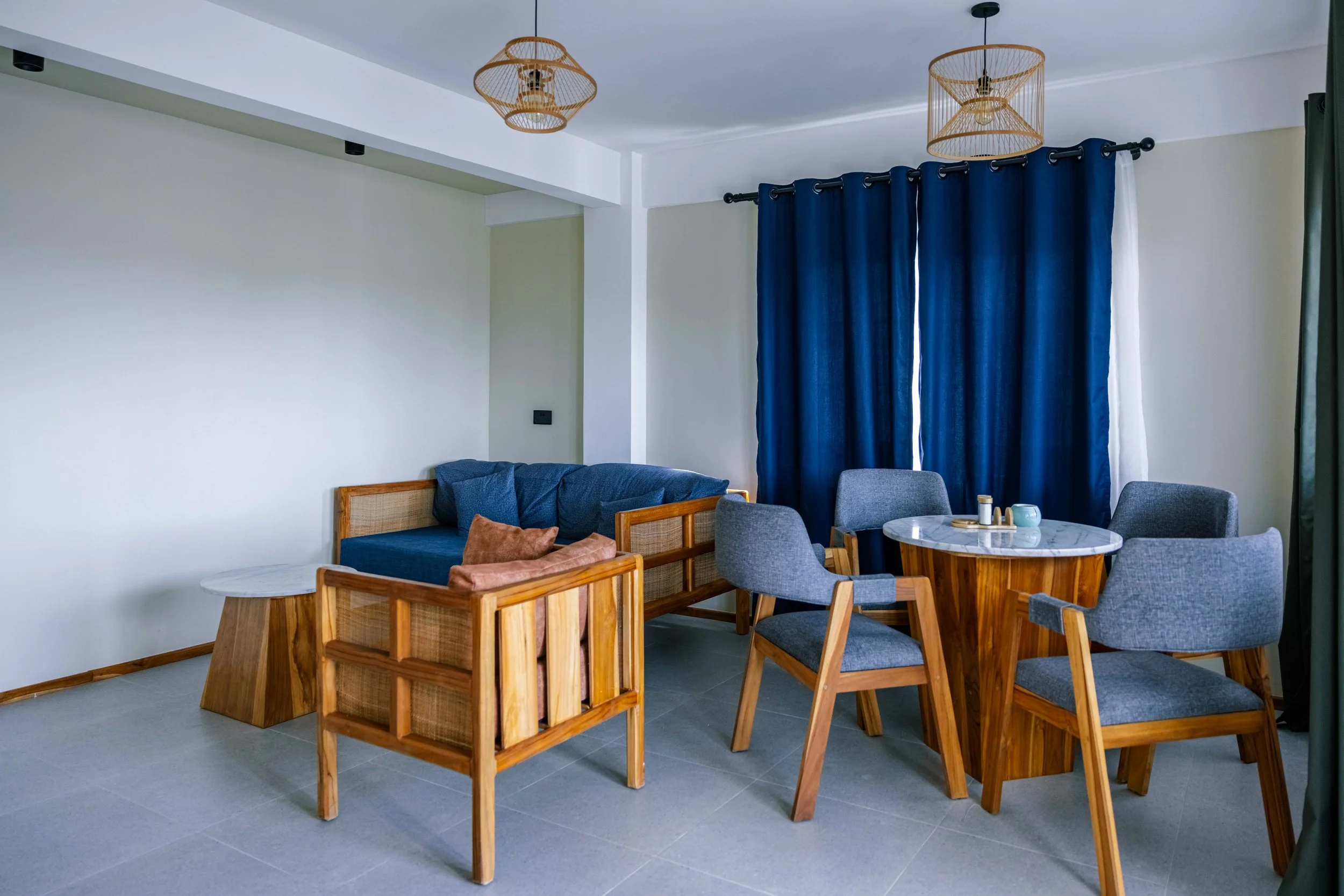 Living or dining area with blue curtains, wooden and fabric chairs, a round marble table, and woven light fixtures.