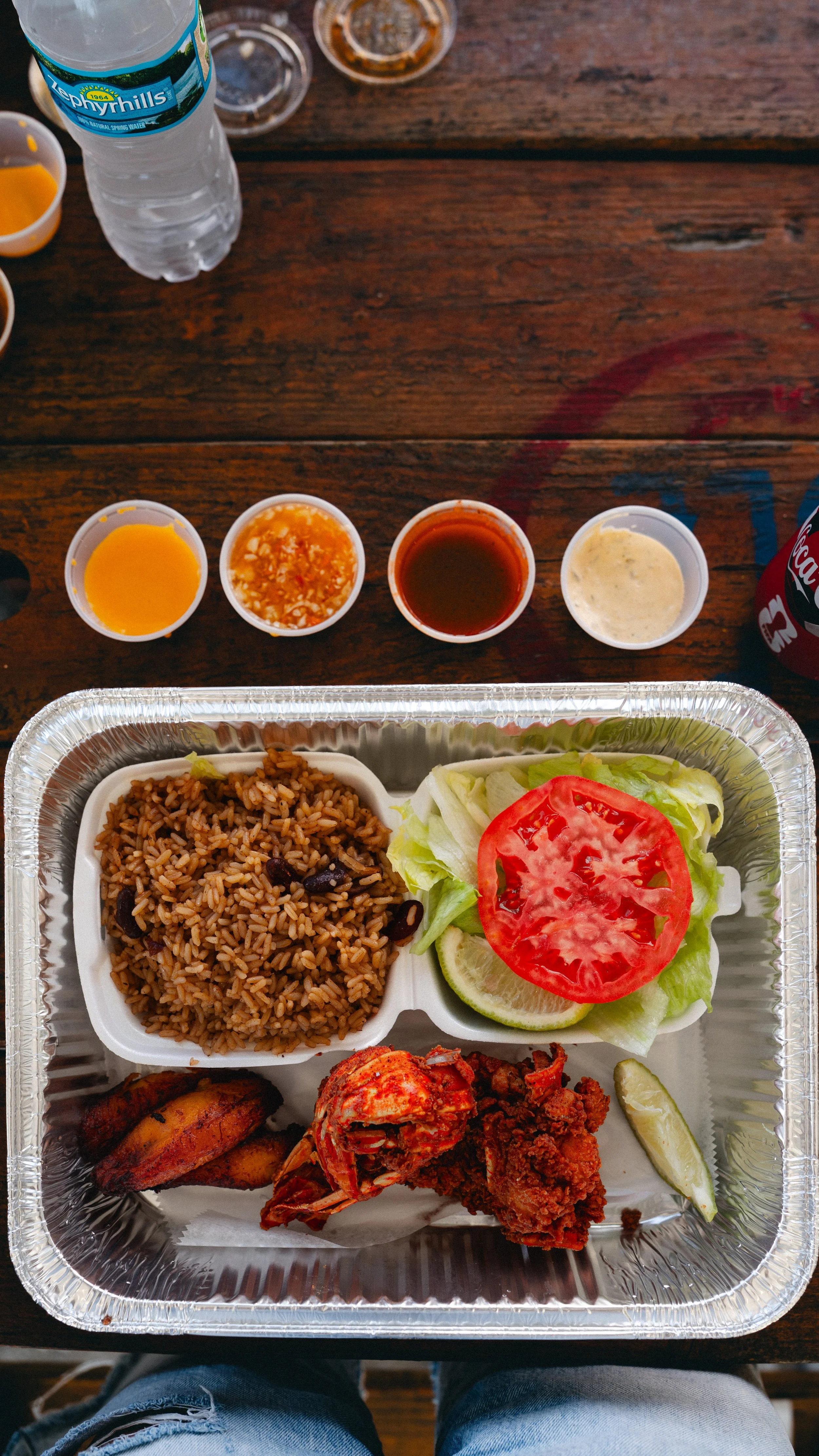 Tri-color rice, fried chicken, tortilla, lettuce with tomato, lemon, and sauces in a takeout tray on a wooden table.