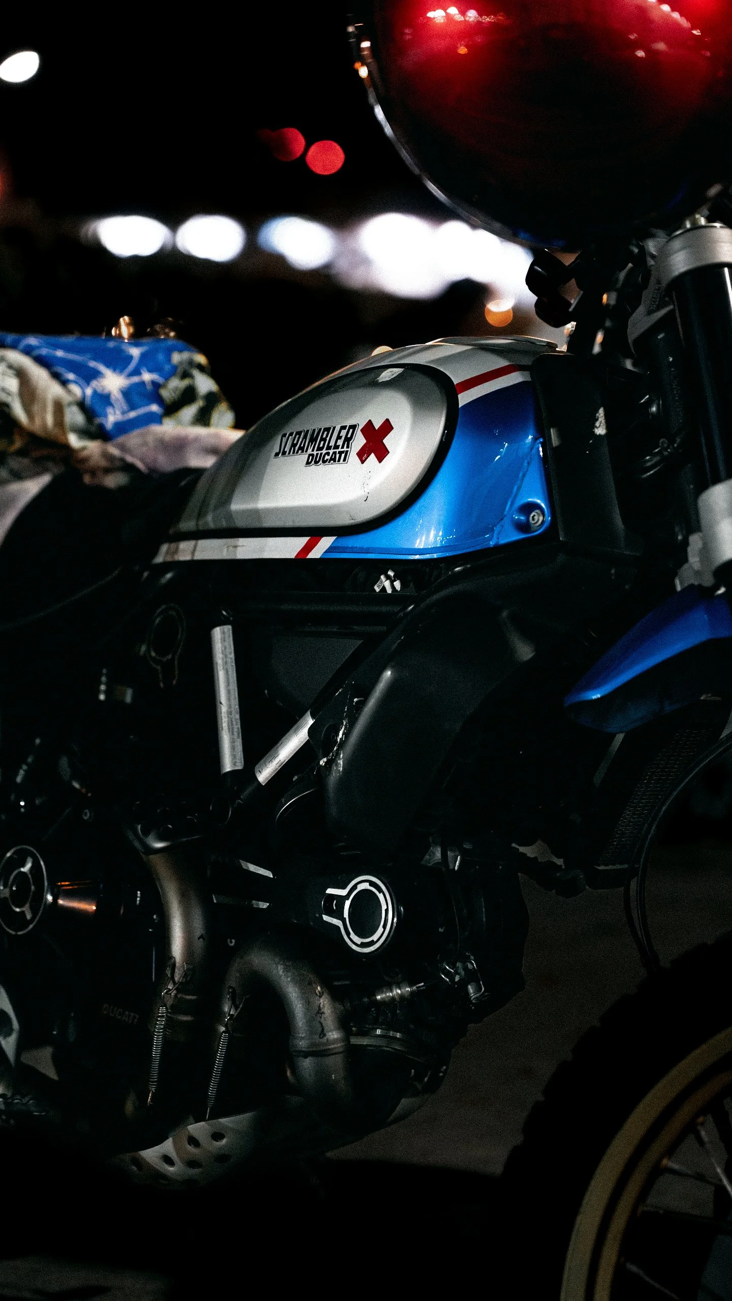 Close-up of a Ducati Scrambler motorcycle with a European blue, white, and red color scheme, parked at night with blurred city lights in the background.