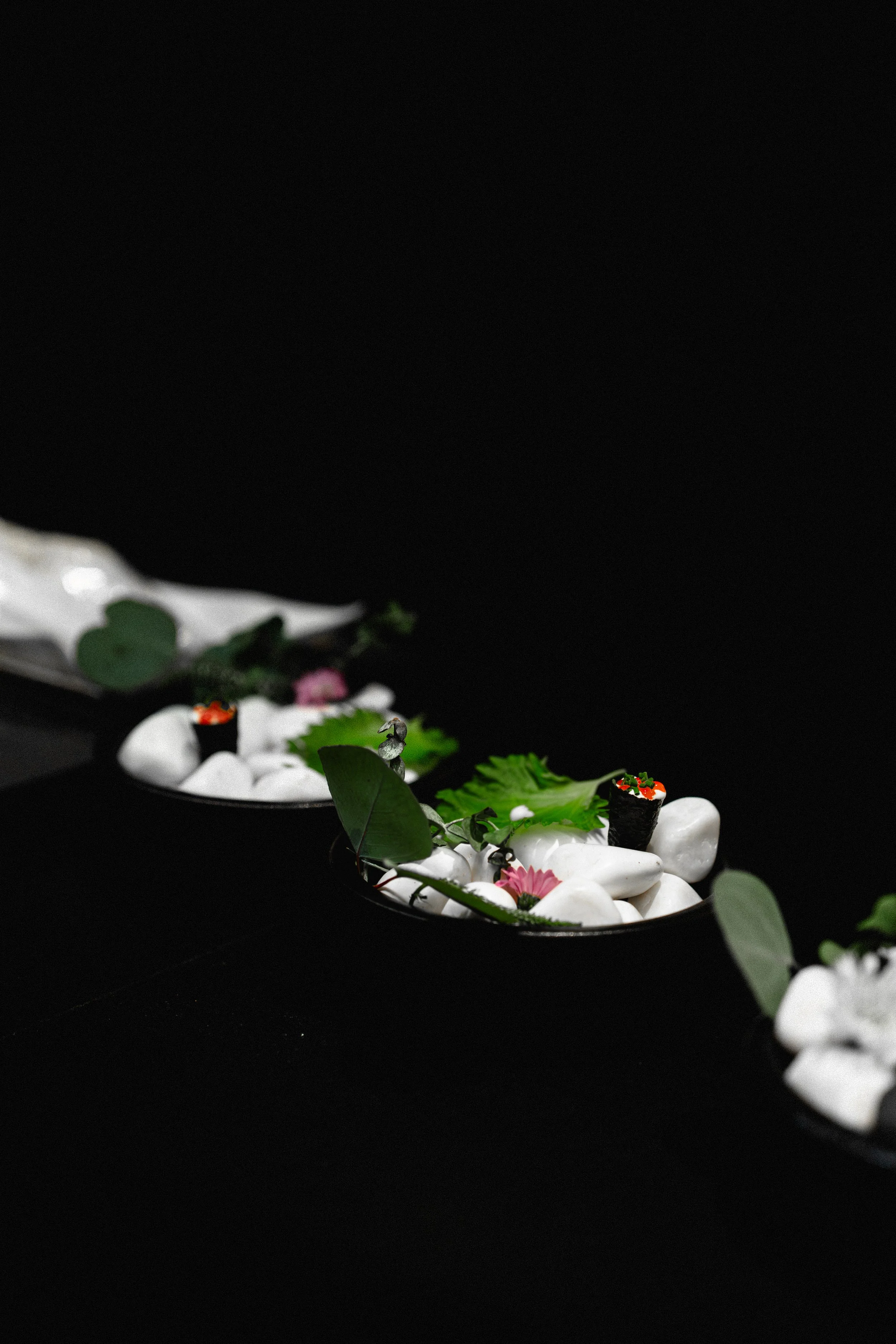 Arrangement of sushi rolls, green leaves, pink flowers, and white stones on black background.