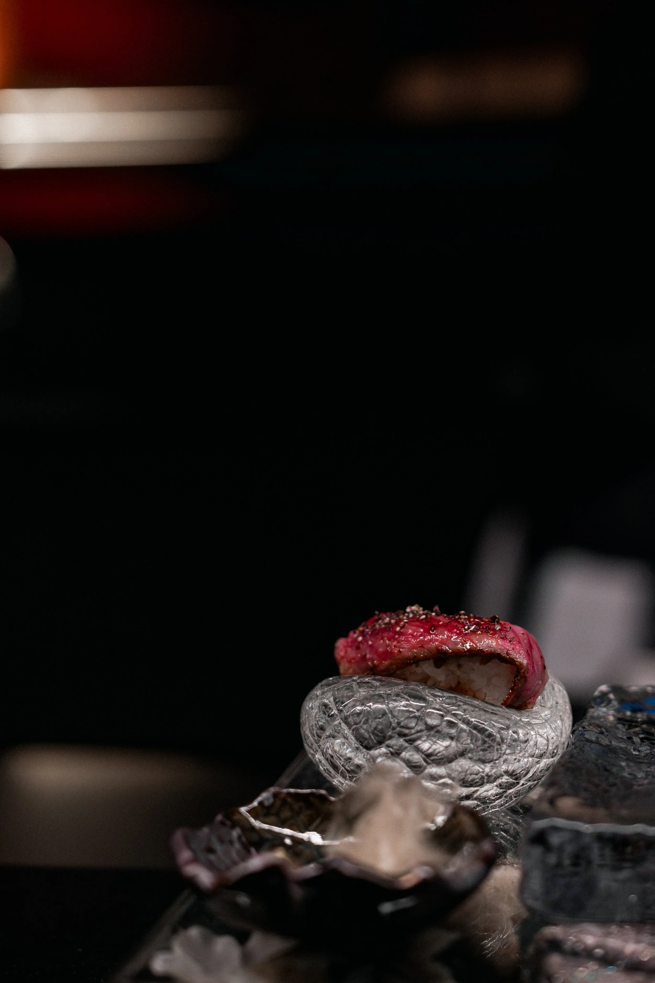 Close-up of a piece of sushi with red topping on small glass bowl, surrounded by ice cubes.