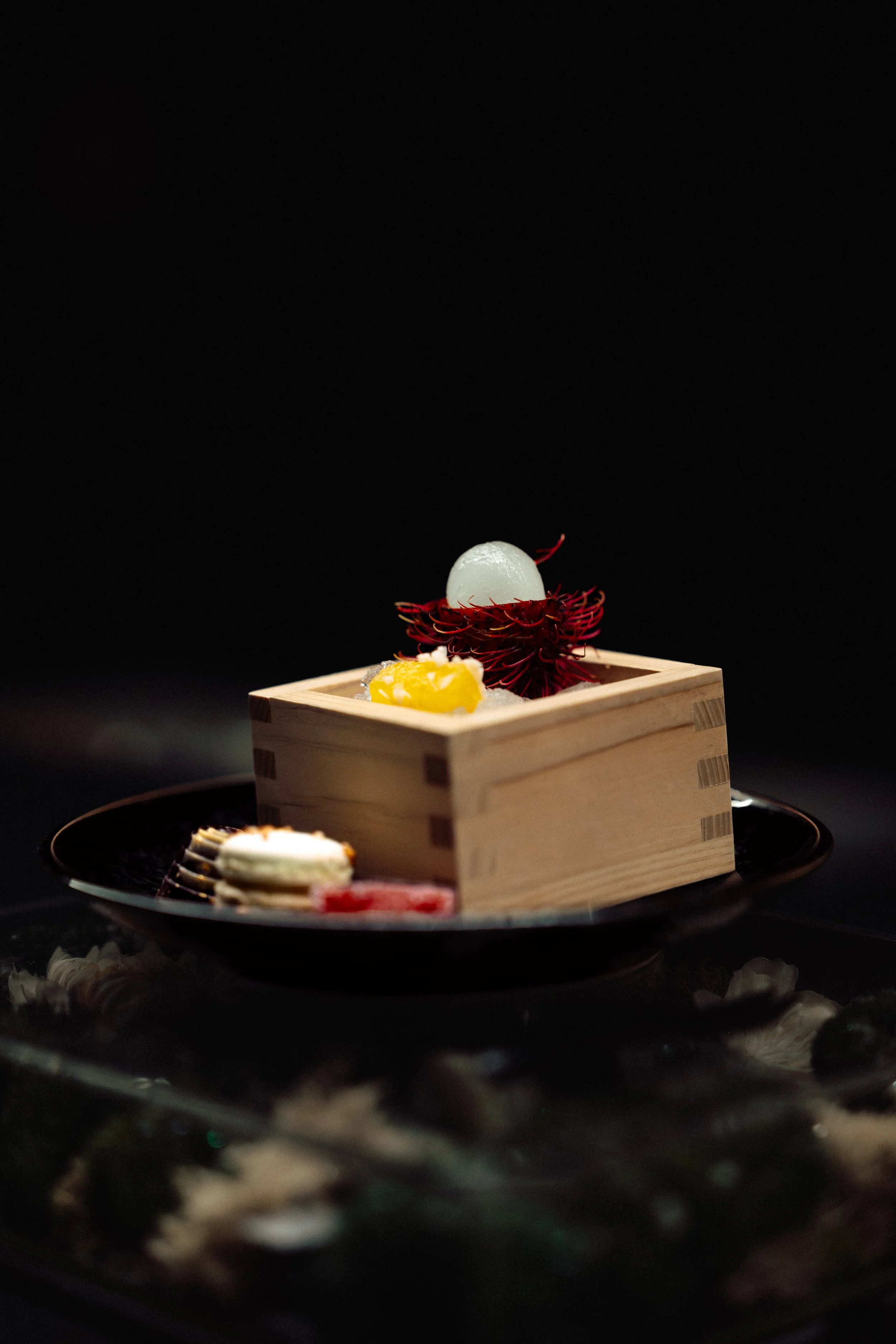 A small sophisticated dessert served on a black plate with a wooden box containing a white spherical treat and a red spiky fruit. Garnished with a yellow lemon-shaped decoration.