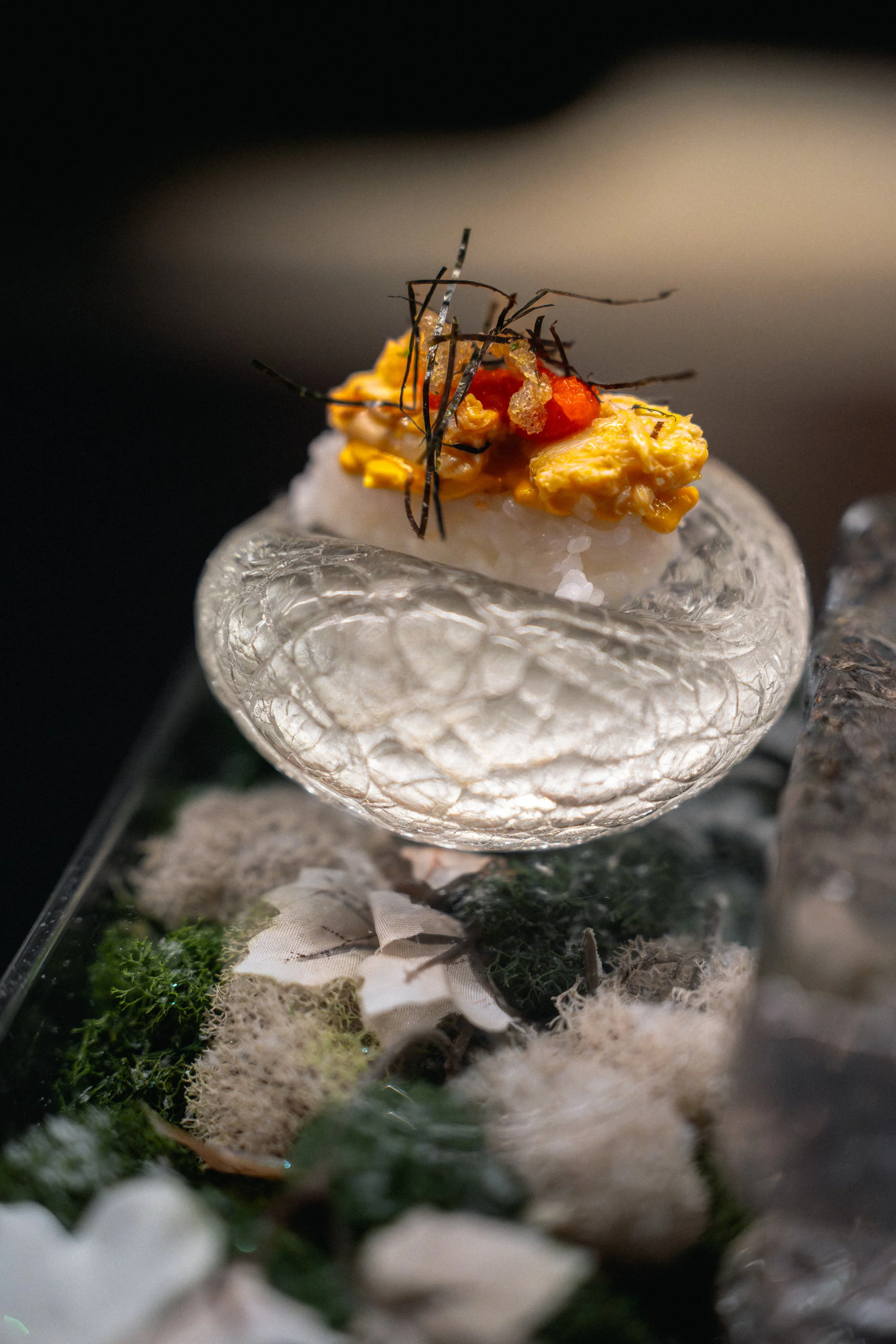 An artistic sushi presentation featuring a piece of sushi topped with scrambled egg, red garnish, and black thin seaweed, placed on a textured glass dish with moss and decorative elements underneath.