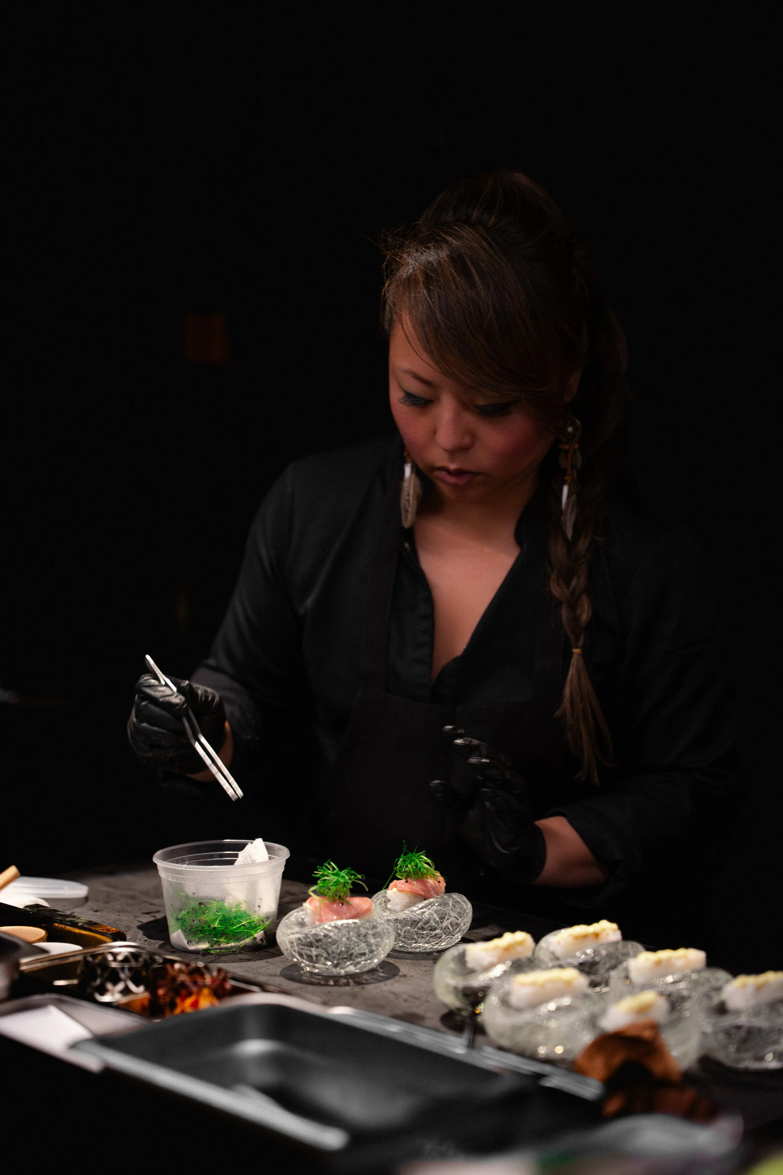 A female chef in black attire and gloves preparing sushi on a dark surface with various ingredients and dishes around.