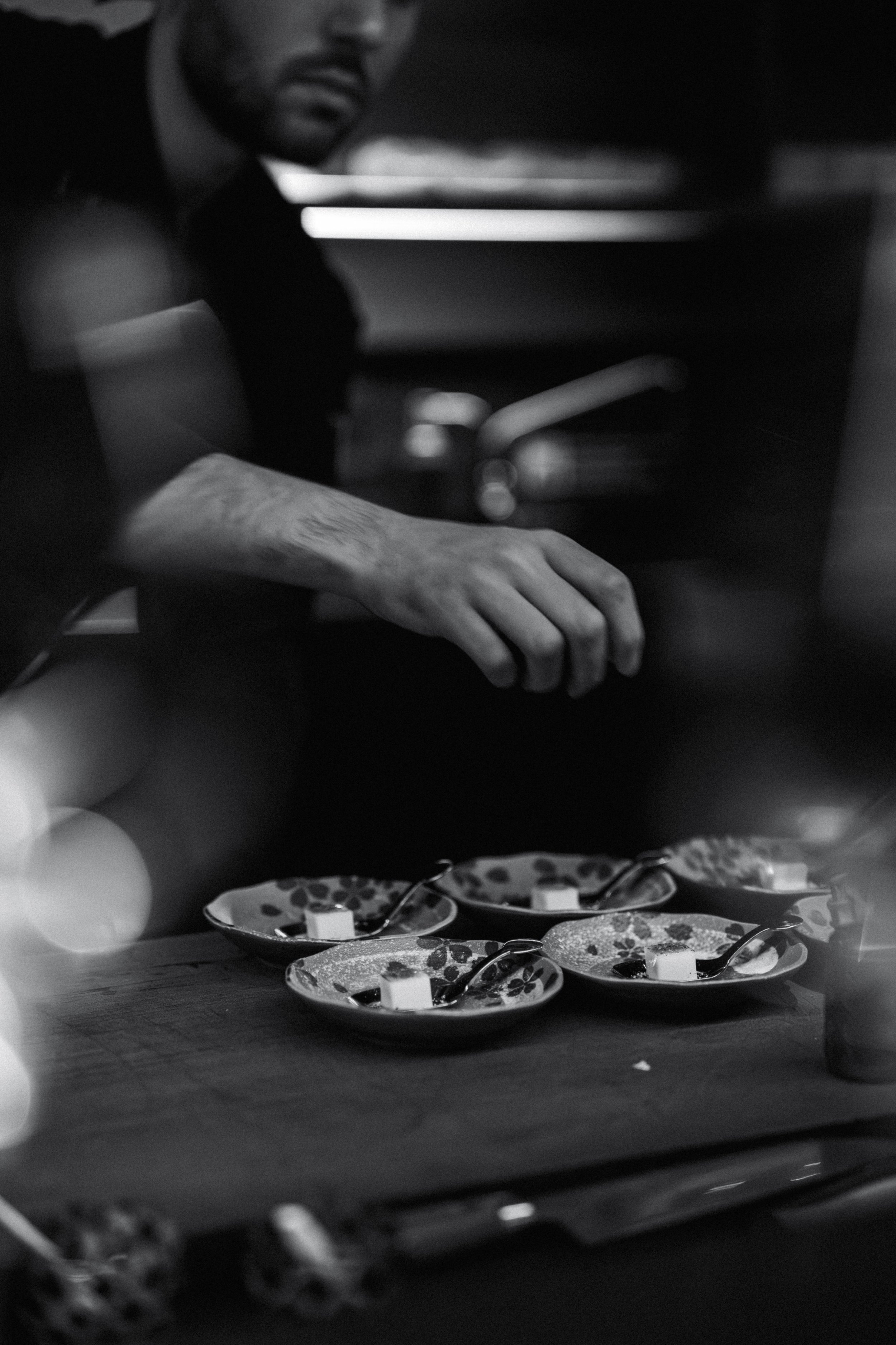 A man in a dark shirt is reaching towards four small dishes with spoons, placed on a table with a rustic cloth. The man has a beard and serious expression, and the image is in black and white with a moody, introspective tone.