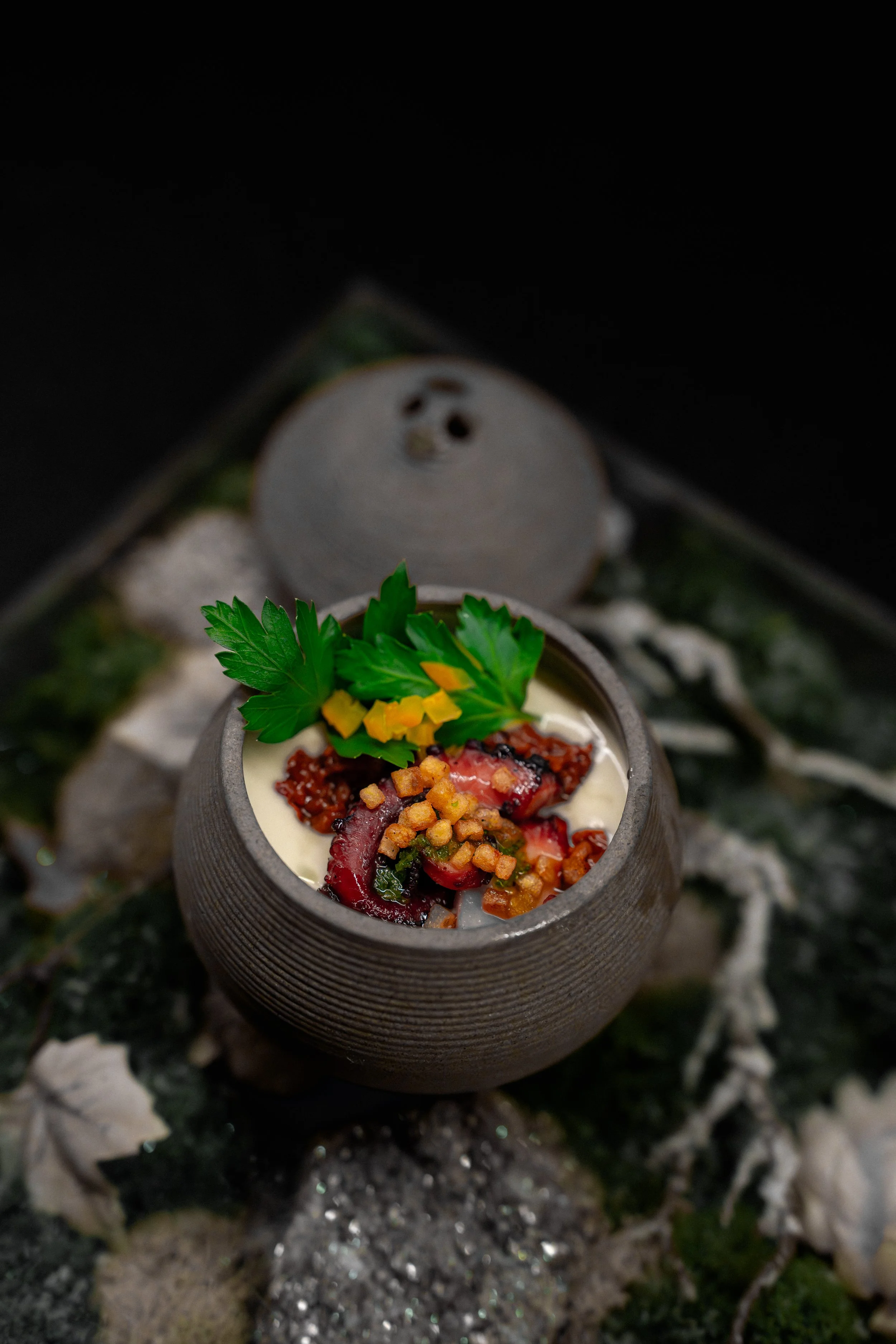 A small stone bowl of gourmet appetizer with diced vegetables, parsley garnish, and a creamy sauce, on a textured surface with dried leaves and moss.