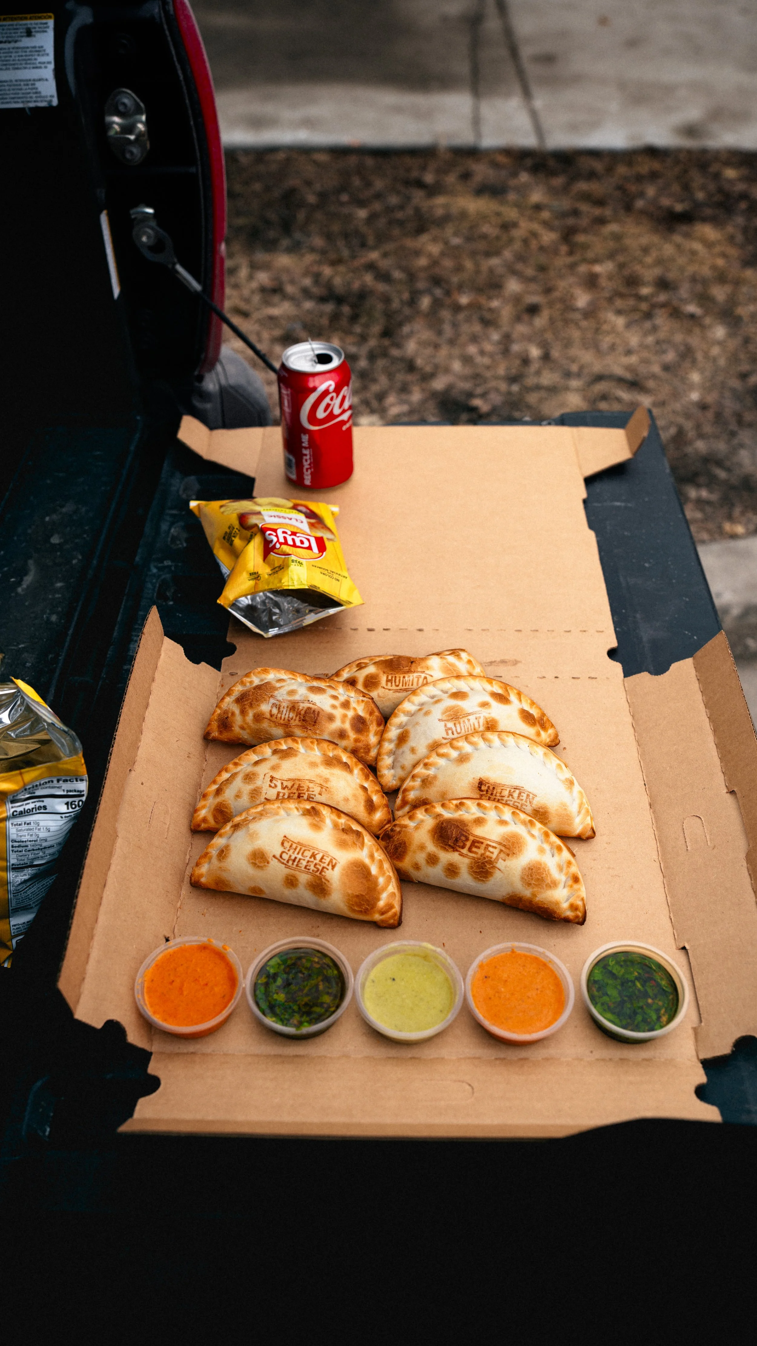 A pizza box with six folded calzones labeled with different fillings, accompanied by five small cups of dipping sauces, a can of Coca-Cola, and a bag of Lay's potato chips.