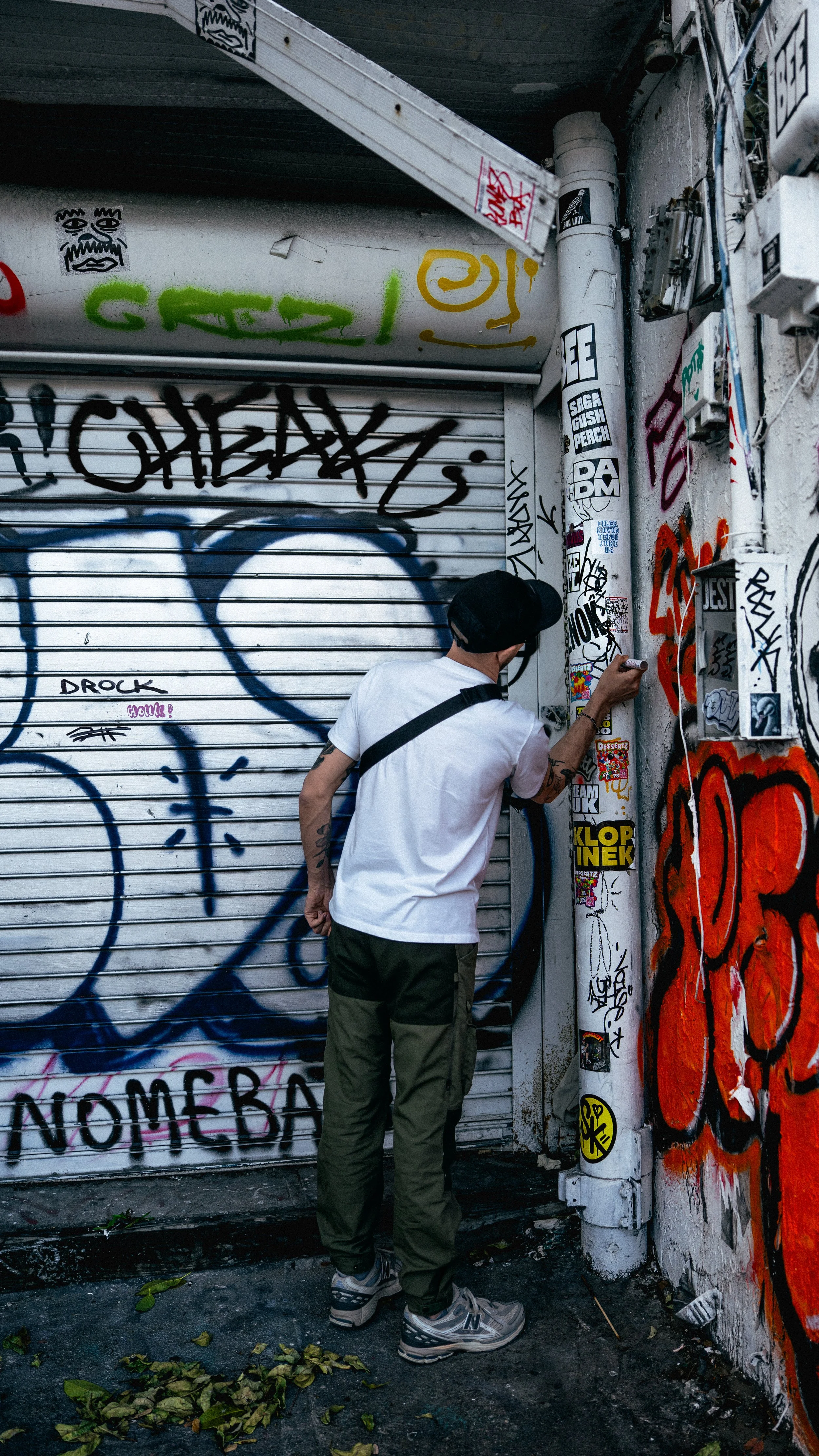 A person wearing a white t-shirt, green pants, sneakers, and a black cap is graffitiing or possibly taking a photo of stickers on a white pole in an alley with graffiti-covered walls and a closed metal shutter.