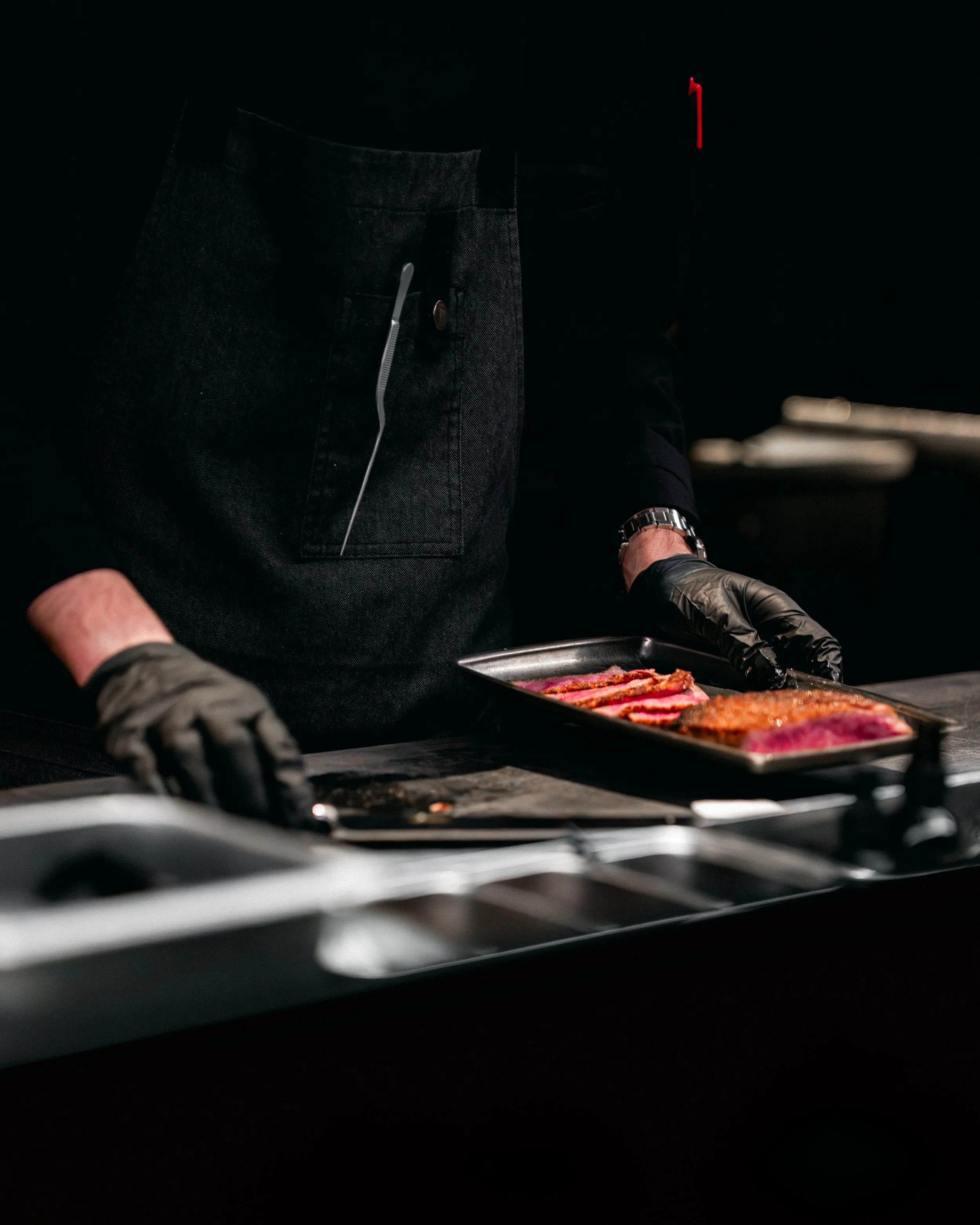 A person wearing black gloves and a black apron is preparing slices of raw steak on a tray indoors with dark lighting.