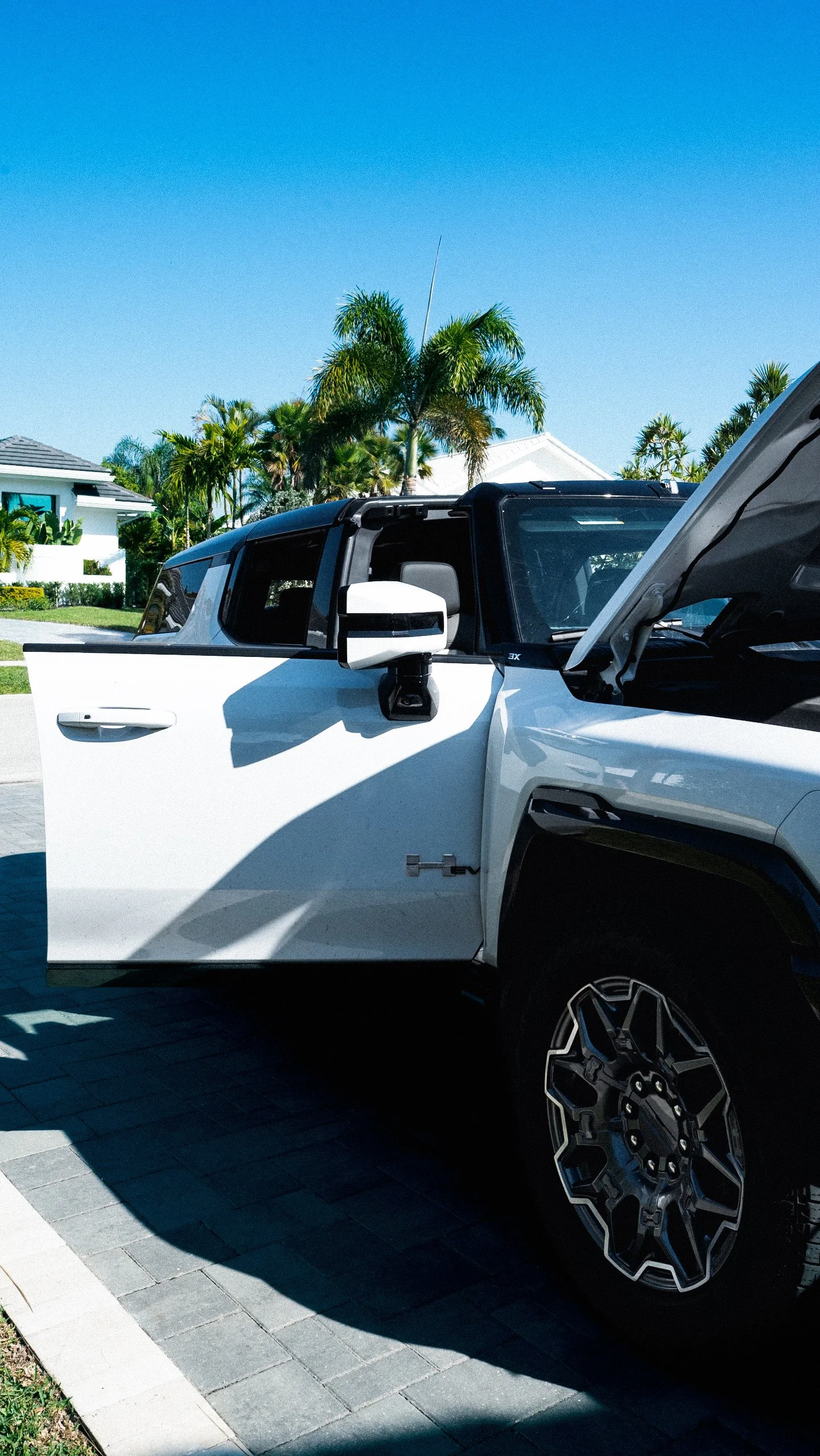 White pickup truck with open hood and driver-side door parked on a paved driveway under a clear blue sky, with palm trees and suburban houses in the background.
