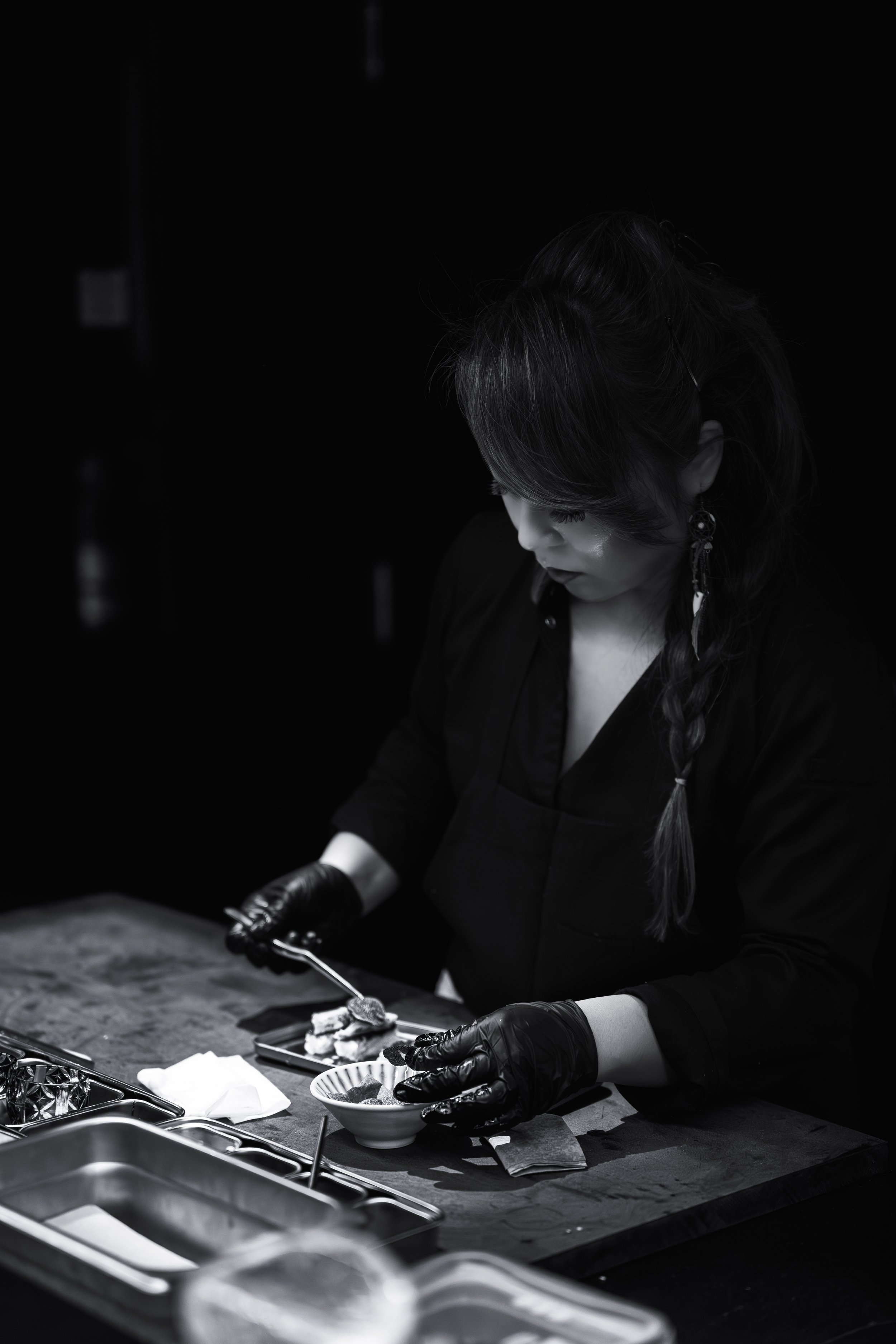 A woman wearing black gloves and earrings is preparing food on a dark counter in a dimly lit setting.