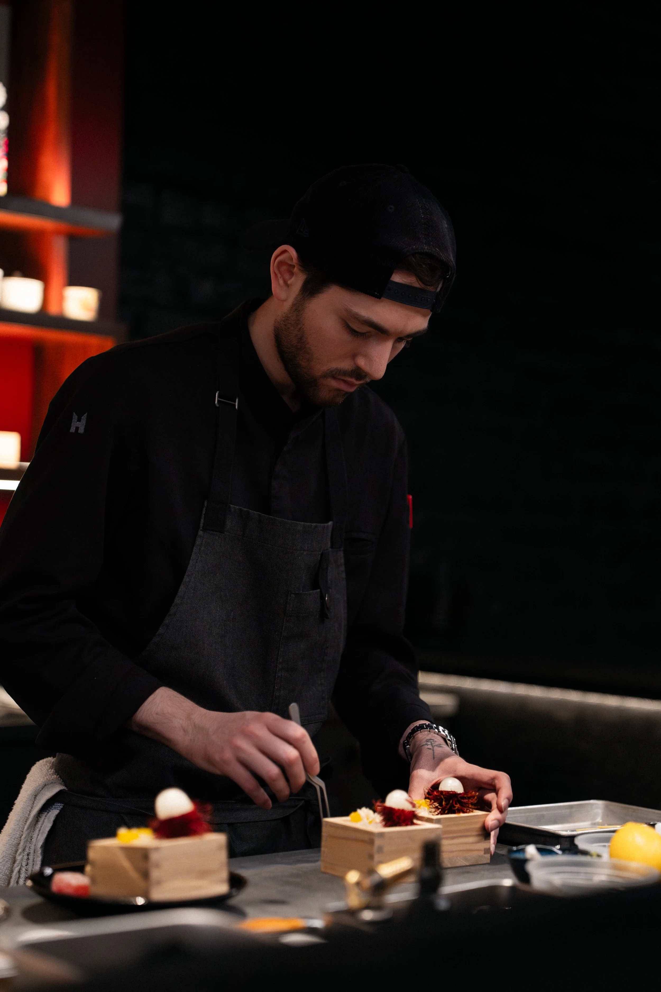 A male chef in black attire and a black cap, focused on plating a dish with tweezers in a professional kitchen with dark background.