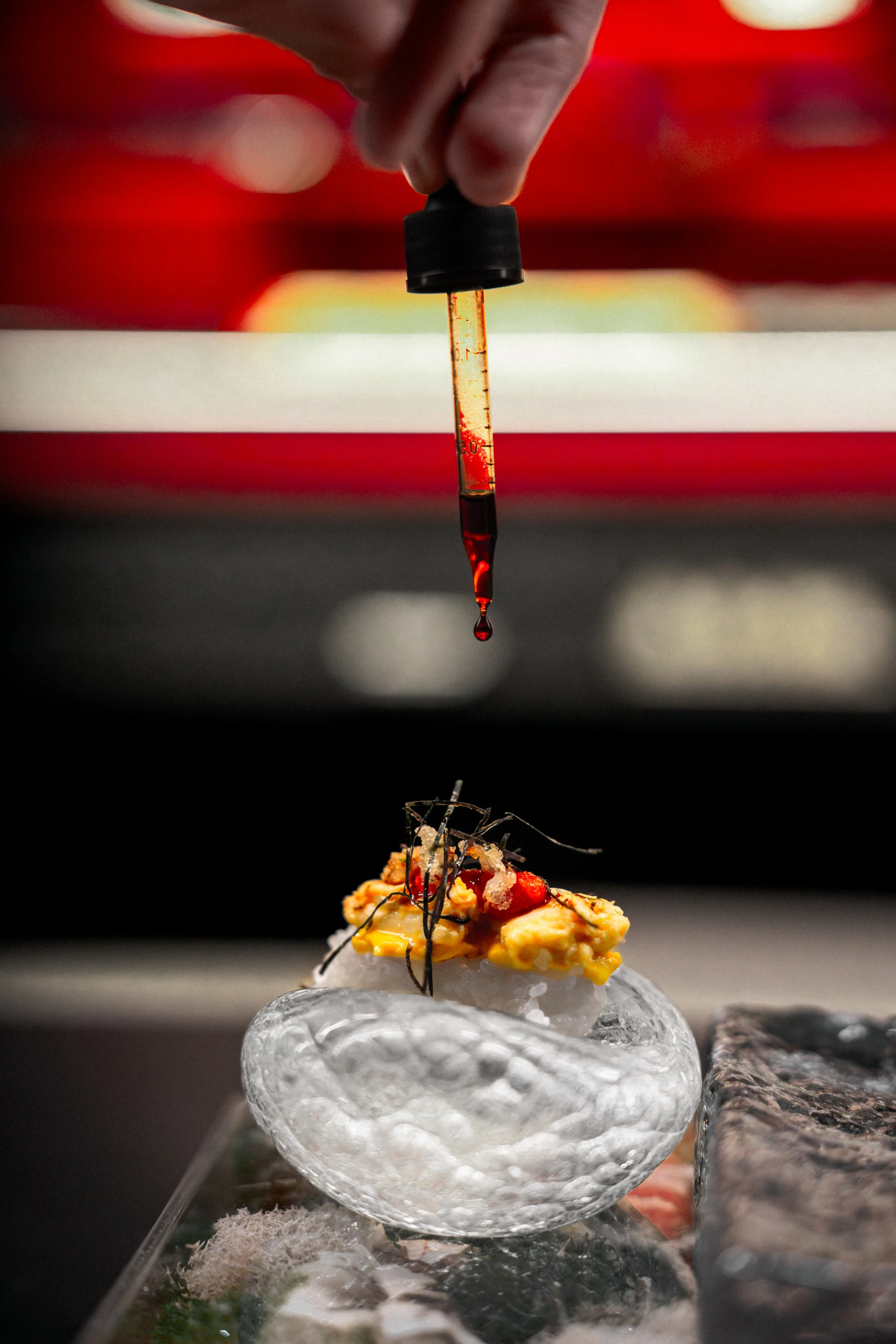 A chef uses a dropper to carefully add red sauce to a piece of sushi topped with egg and garnished with black shredded seaweed on a glass plate.