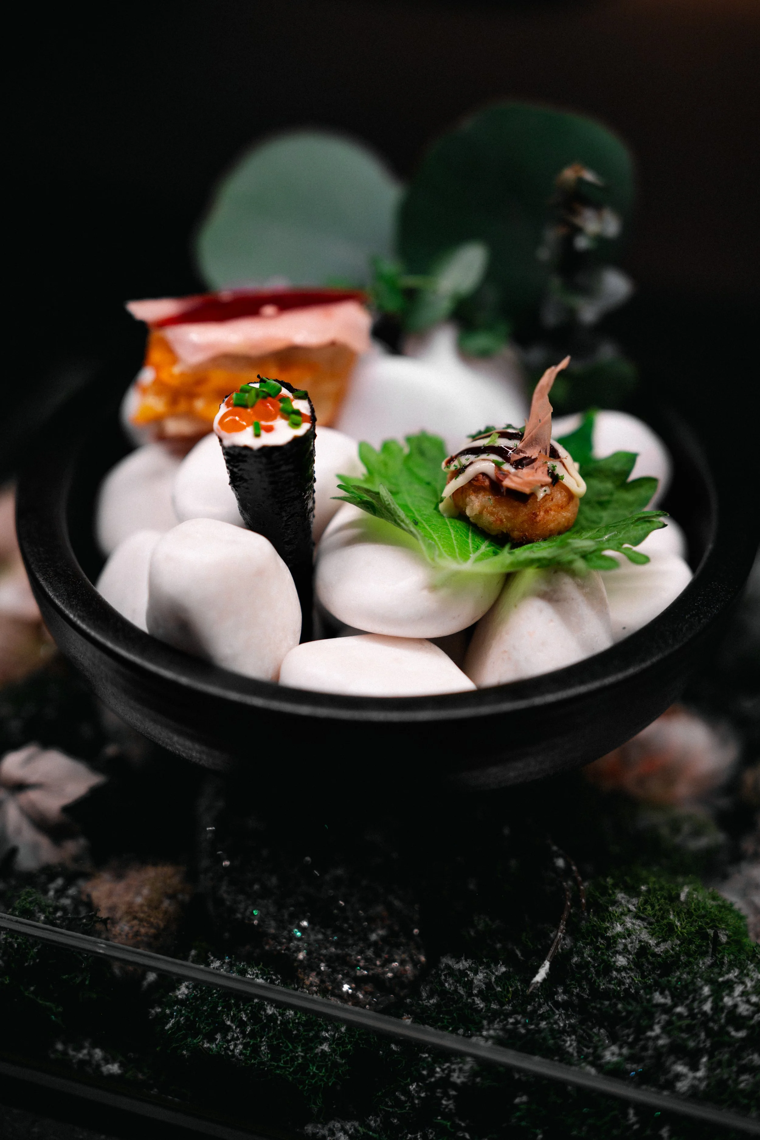 Assorted gourmet appetizers, including sushi roll, fried croquette, and small sandwich, served on white stones with green leaves in a black bowl.