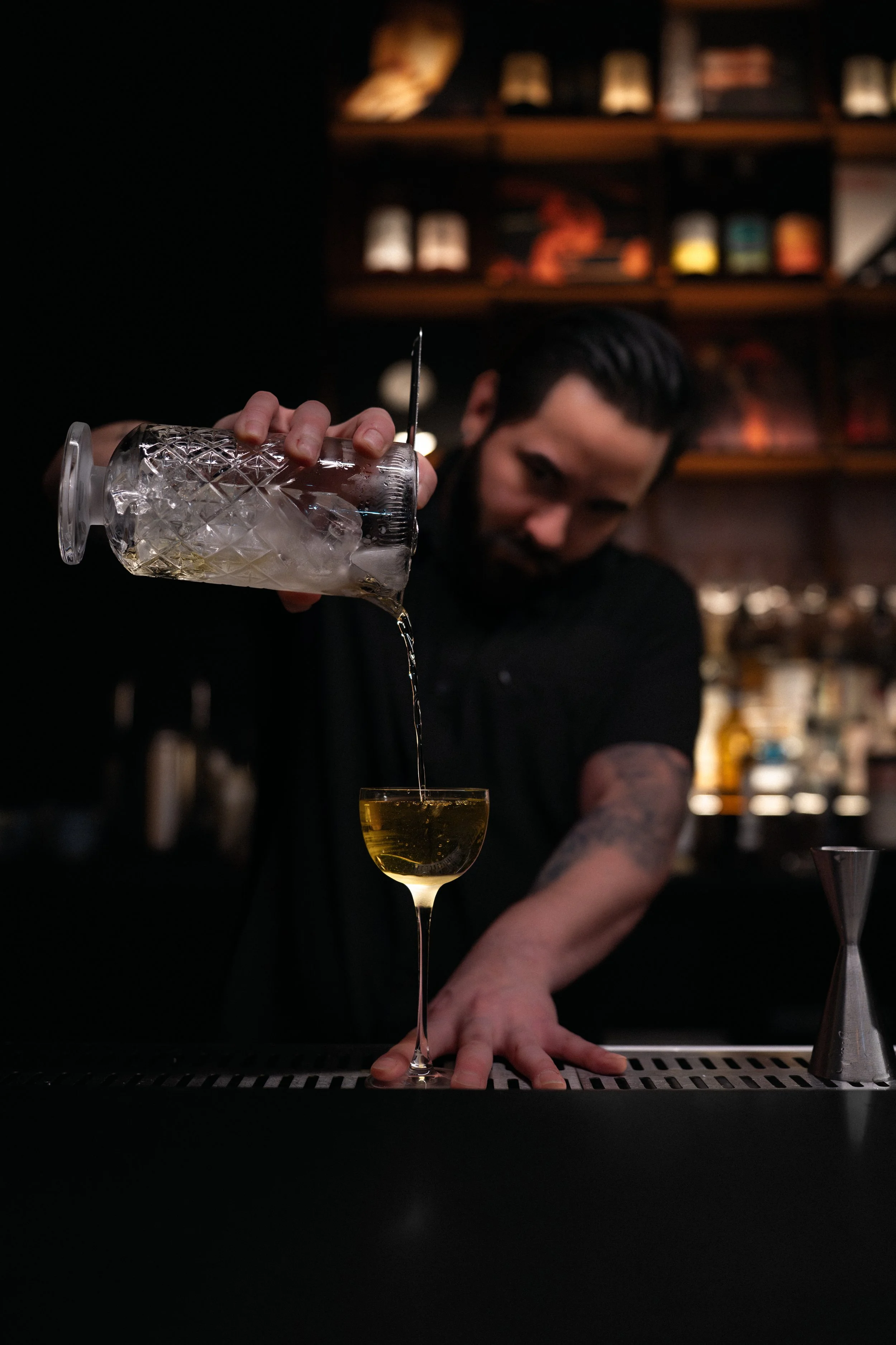 Bartender pouring yellow cocktail into a coupe glass in a dimly lit bar.