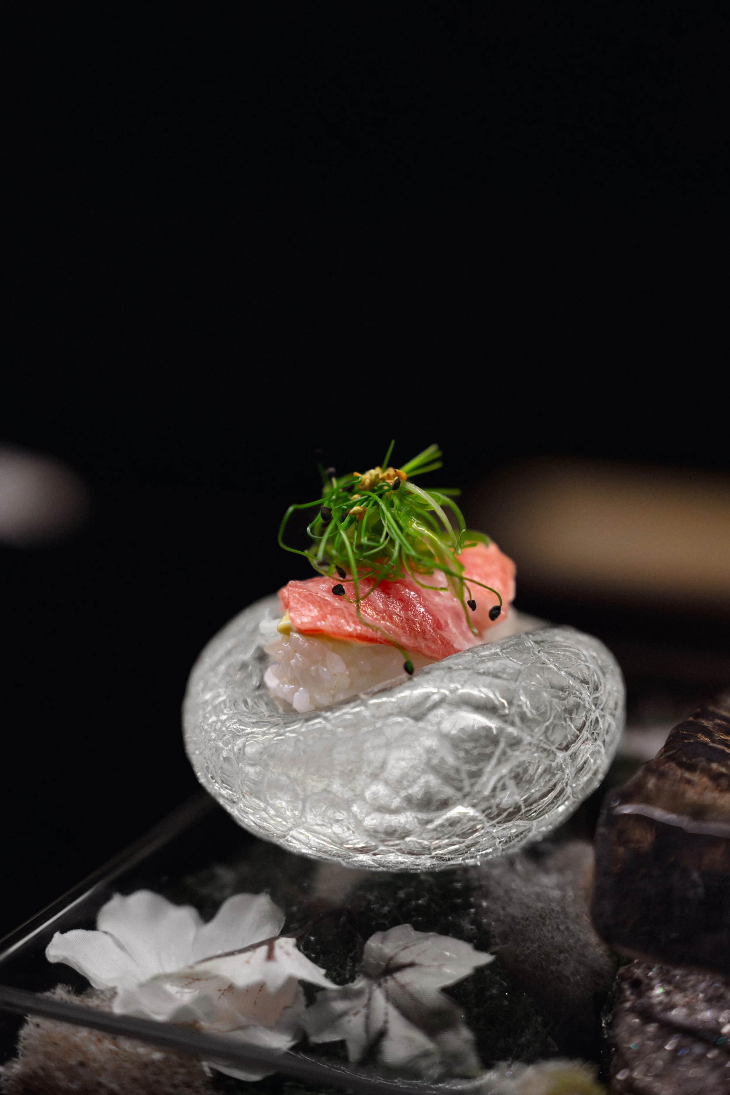 Close-up of a glass dish with sushi topped with a slice of raw fish, microgreens, and garnished with black sesame seeds, set against a black background.