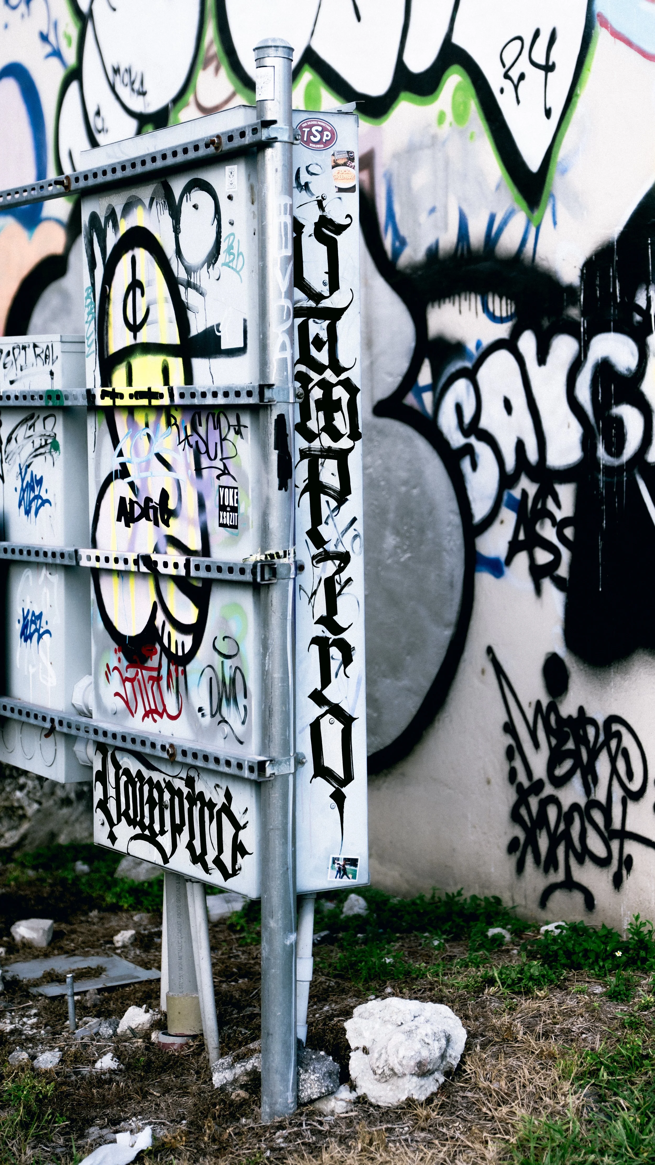 A metal electrical box covered in various colorful graffiti and stickers, with a large black graffiti face painted on the wall behind it.