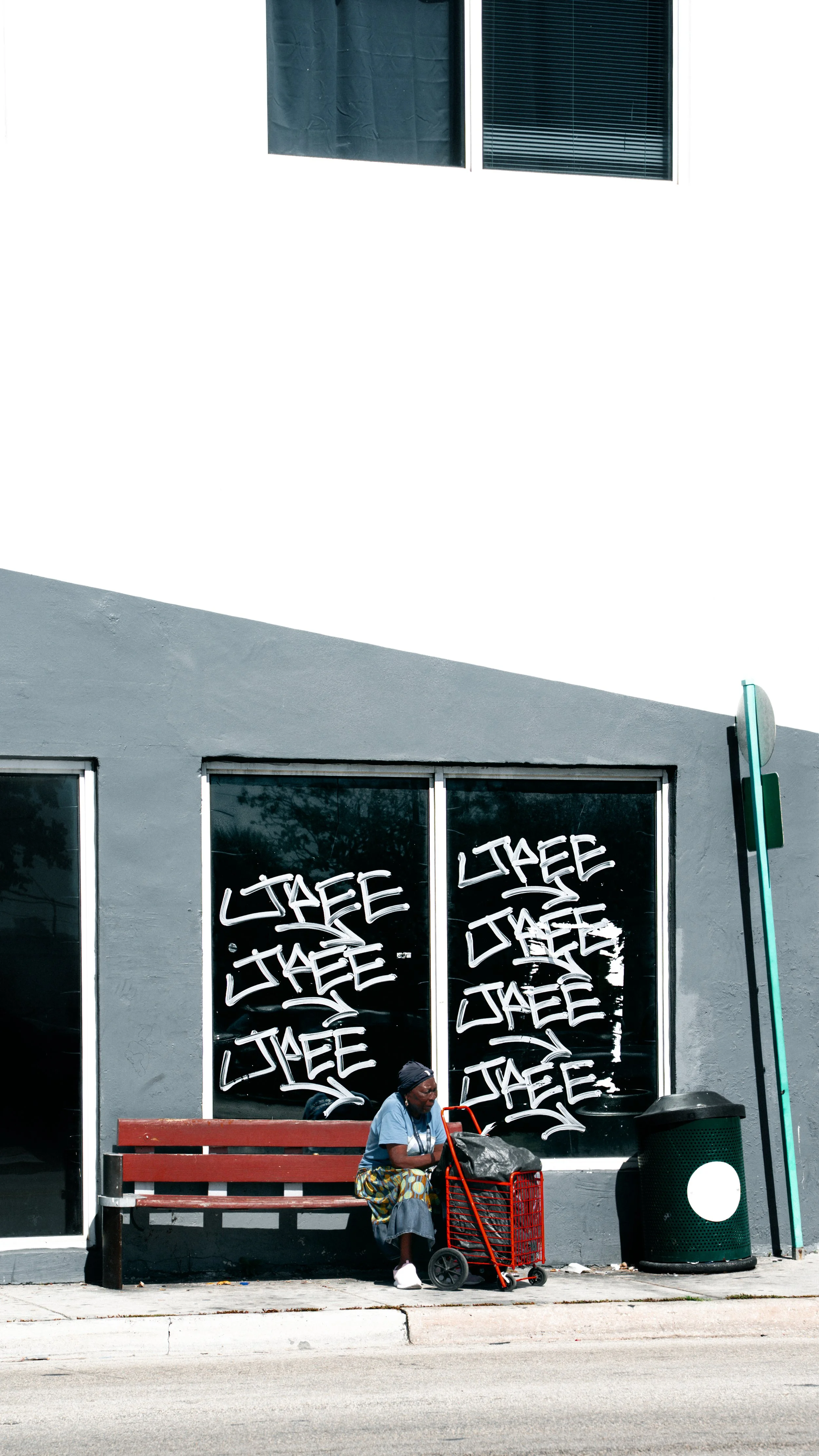 A woman sitting on a red bench outside a building with graffiti-covered windows, next to a trash can and a teal pole, during daytime.