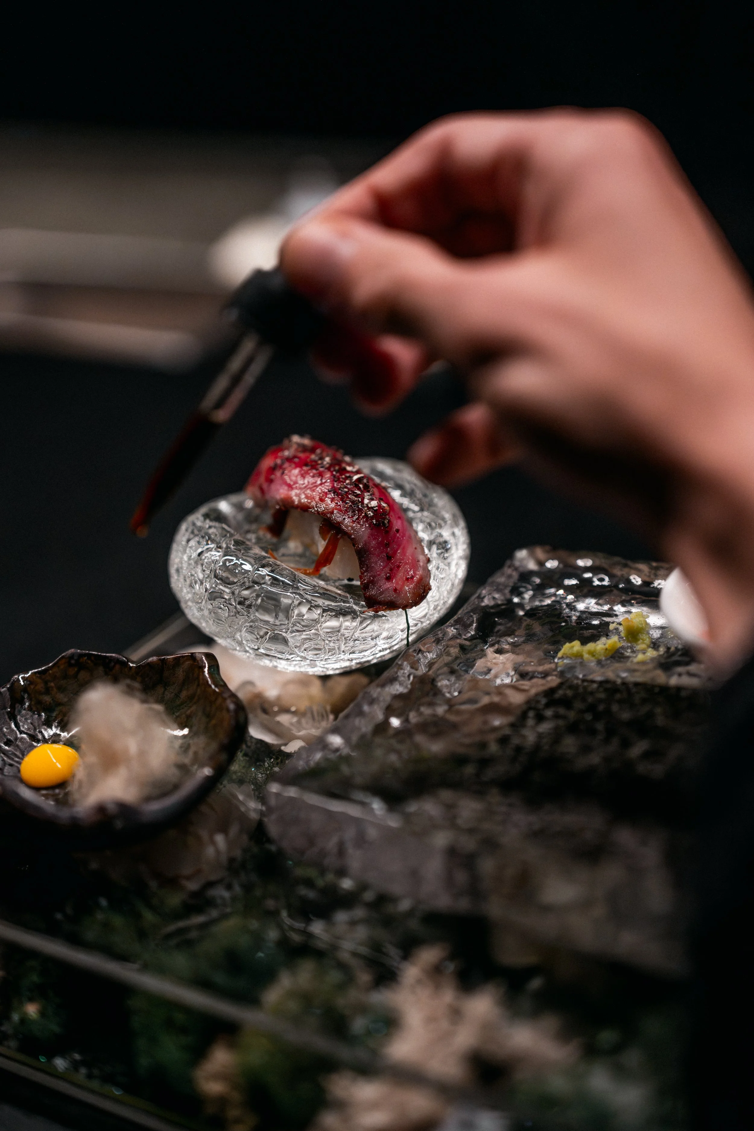 A chef or cook uses tweezers to place a piece of roasted or grilled meat on a textured glass plate, with other small dishes and green garnish on the dark surface.