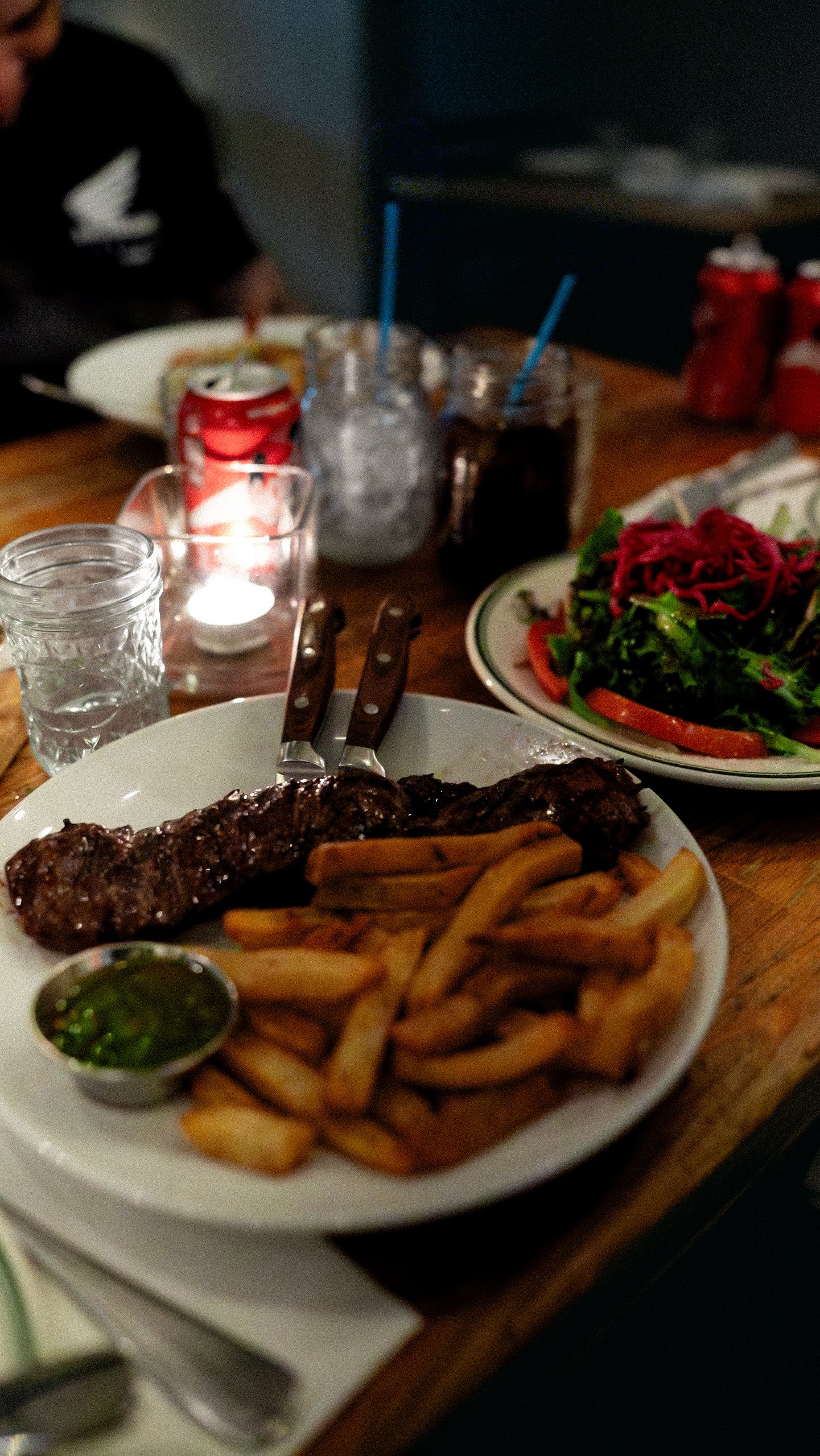 Plate of grilled steak, French fries, and green dipping sauce, with side salad of greens and tomatoes, on a wooden table at a restaurant.