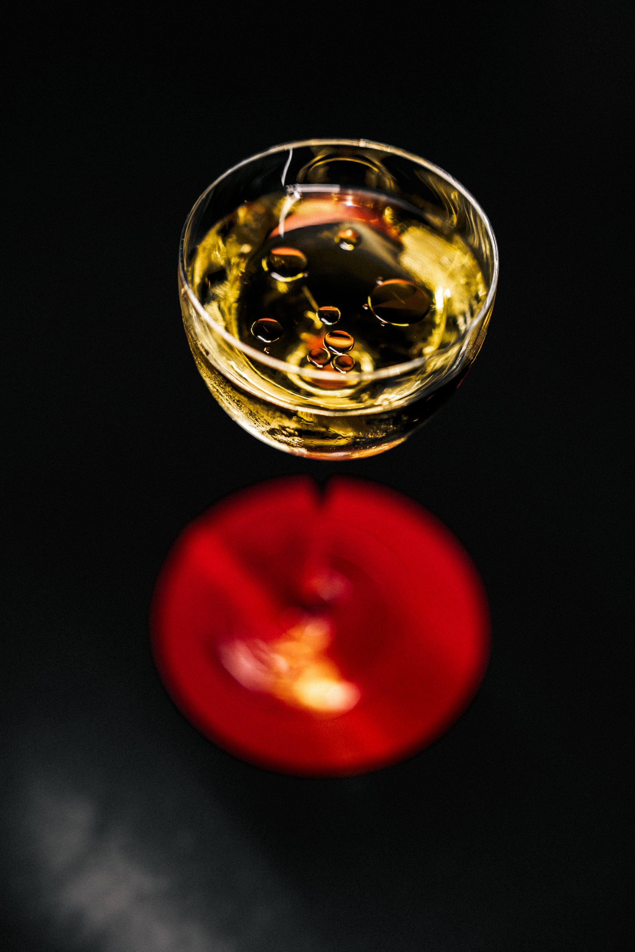 A top-down view of a glass of white wine on a black surface with a red apple placed beneath the glass, causing the apple to appear blurred and distorted due to refraction.