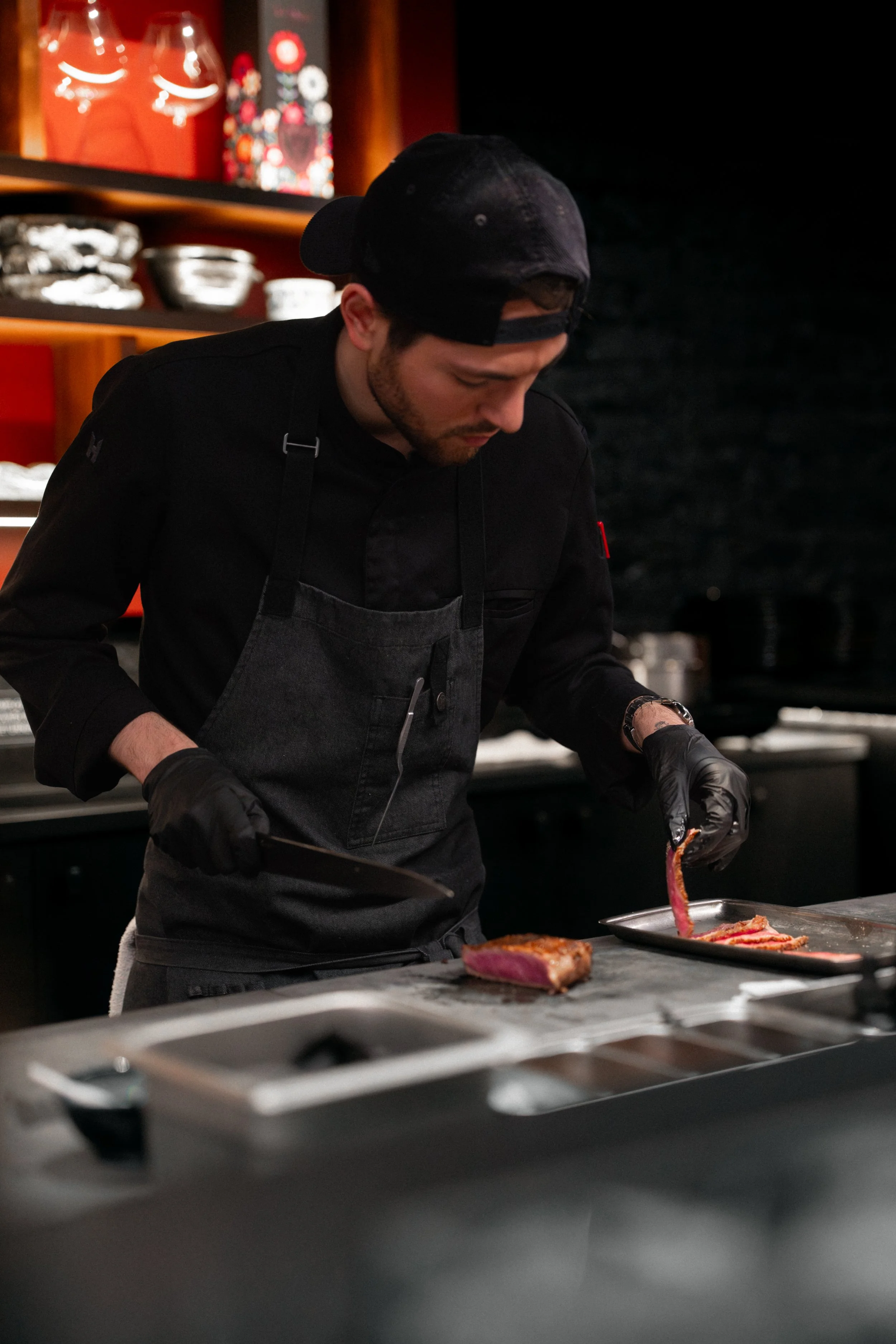 A chef wearing a black shirt, black apron, black gloves, and a backwards black cap prepares a cut of grilled meat in a professional kitchen.