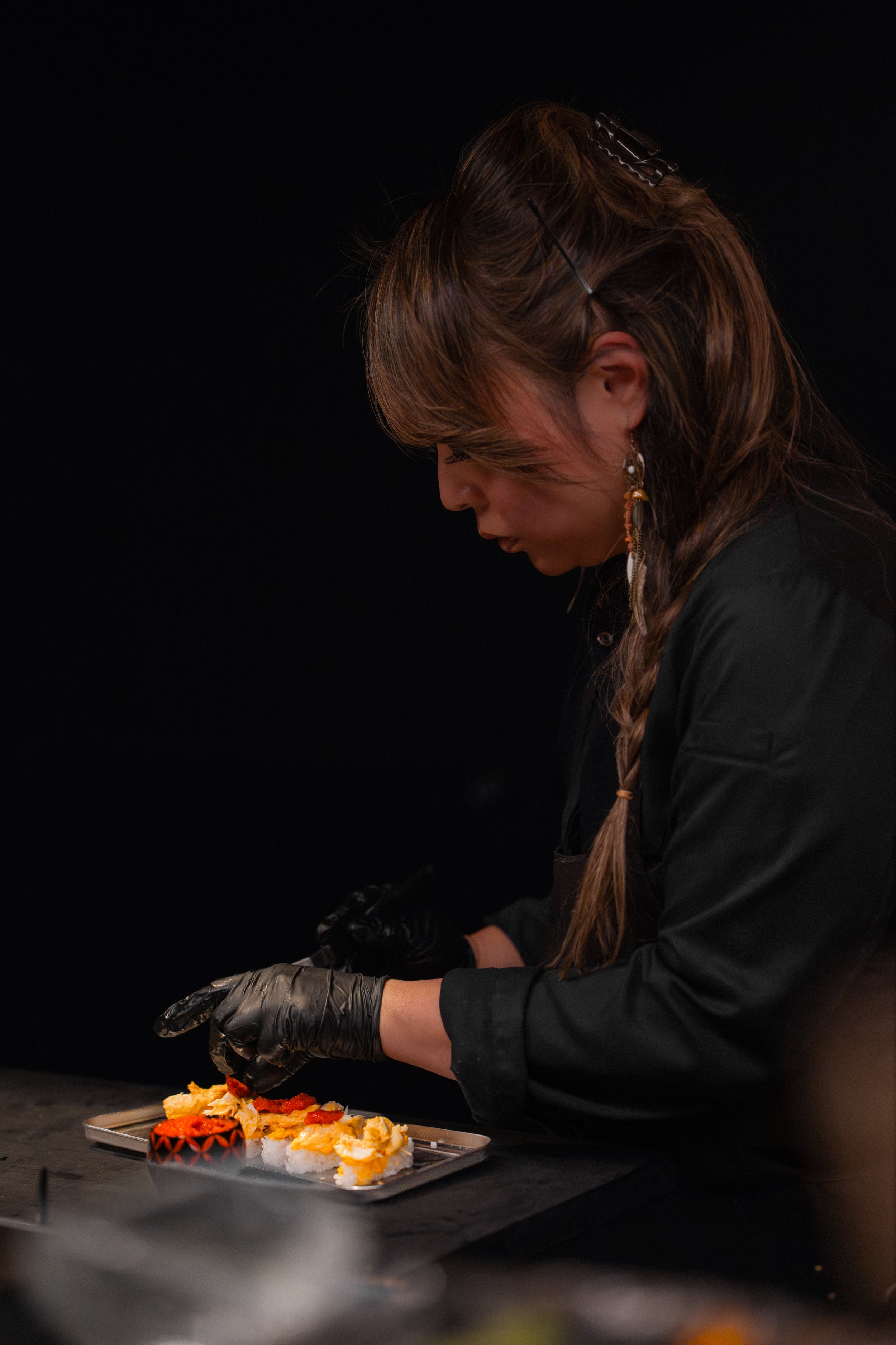 A woman with braided hair, wearing gloves and earrings, carefully preparing sushi on a tray against a dark background.
