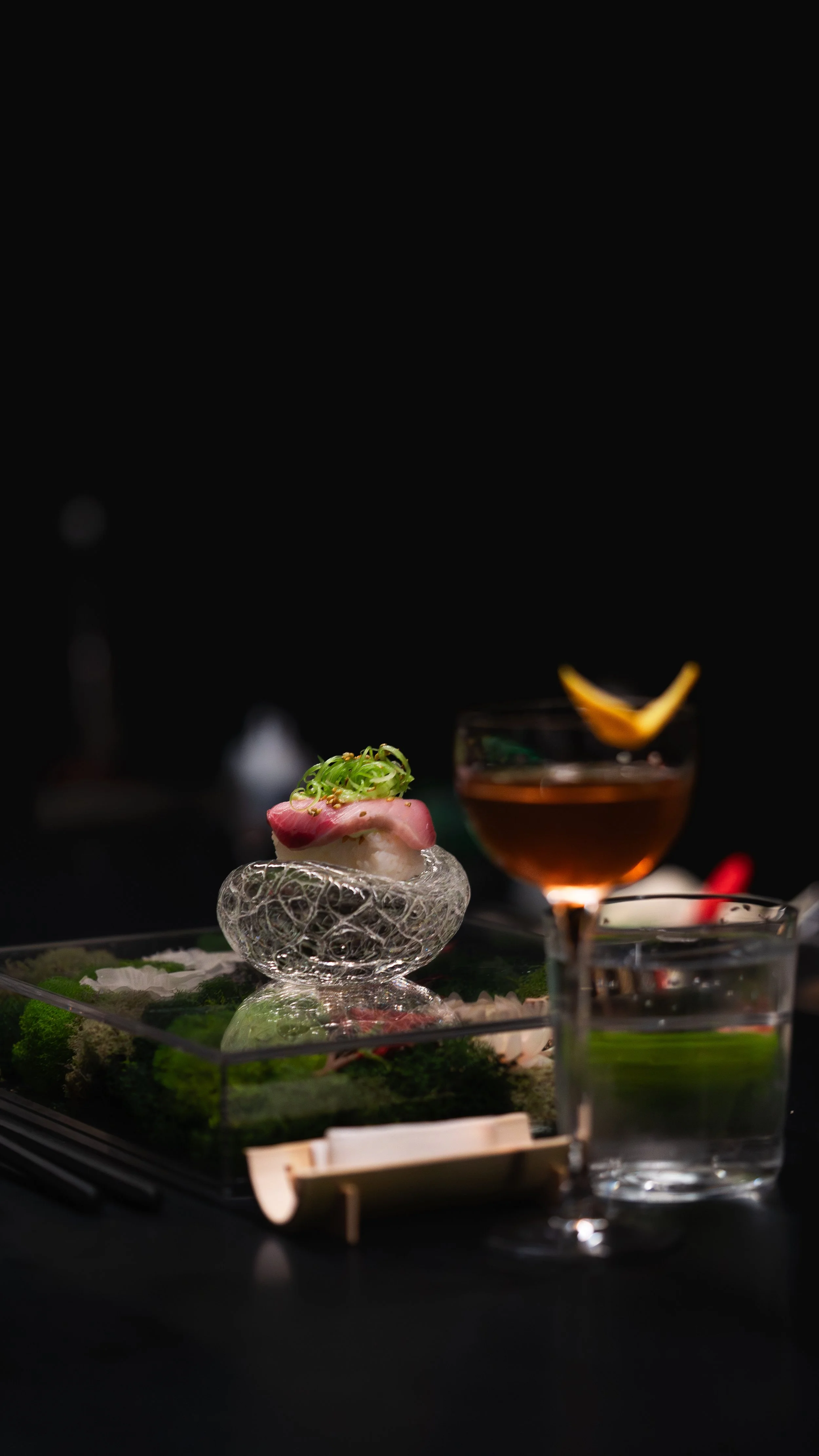 A fancy dish with a piece of fish topped with green onions, served on a glass sculpture, accompanied by a glass of brown beverage with a lemon wedge and a glass of water on a dark table.
