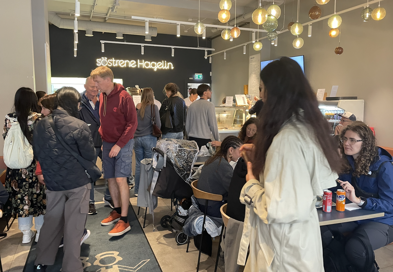 People standing and sitting inside a busy café or restaurant. The background features a black wall with the illuminated sign 'söstrene Hagelin'. Some customers are ordering at the counter, while others are seated and waiting. The space has modern decor with colorful hanging glass orbs from the ceiling.