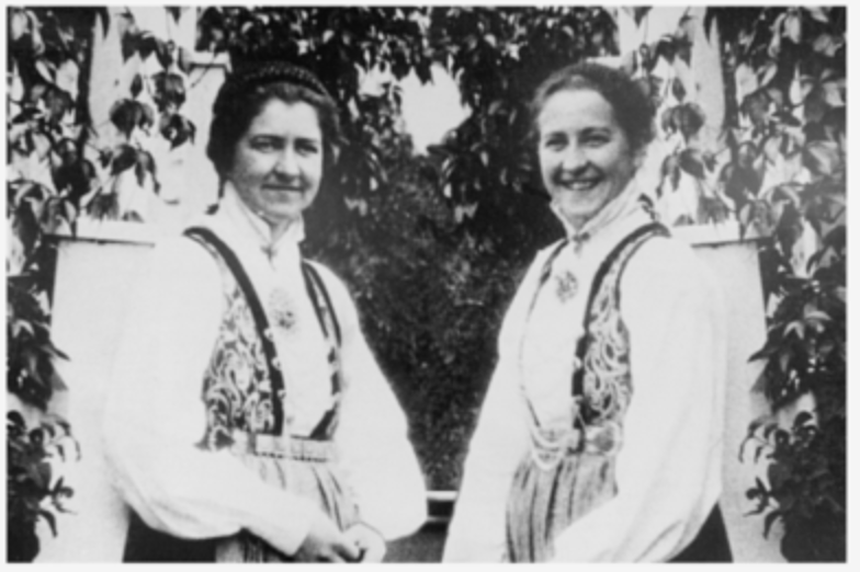 Two women in traditional folk attire standing outdoors among trees, smiling at the camera.
