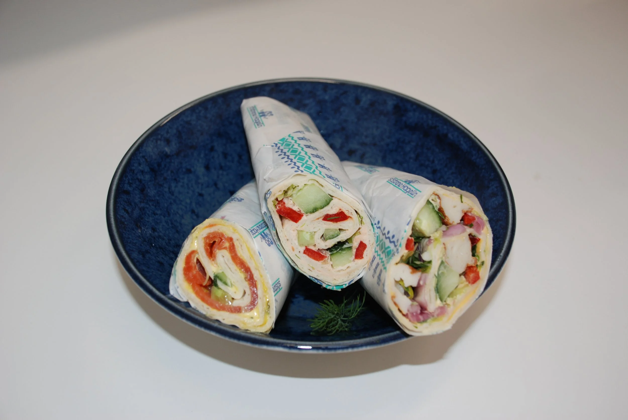 A dark blue bowl filled with sliced vegetable wraps, showcasing ingredients like cucumbers, red peppers, and other vegetables inside a creamy wrap. The bowl is placed on a white surface.