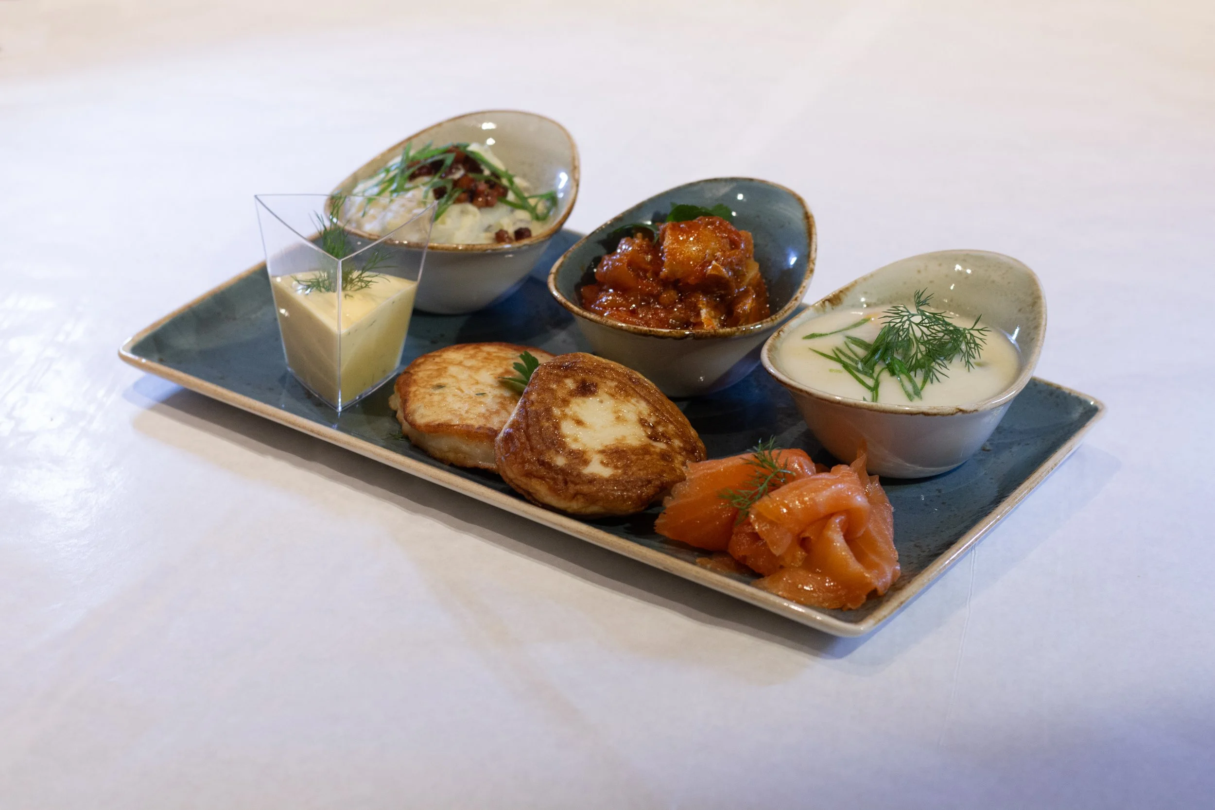 An elegant presentation of Indian cuisine on a rectangular ceramic plate, featuring three small bowls with different dishes, a grilled patty, and a side of smoked salmon, garnished with herbs.