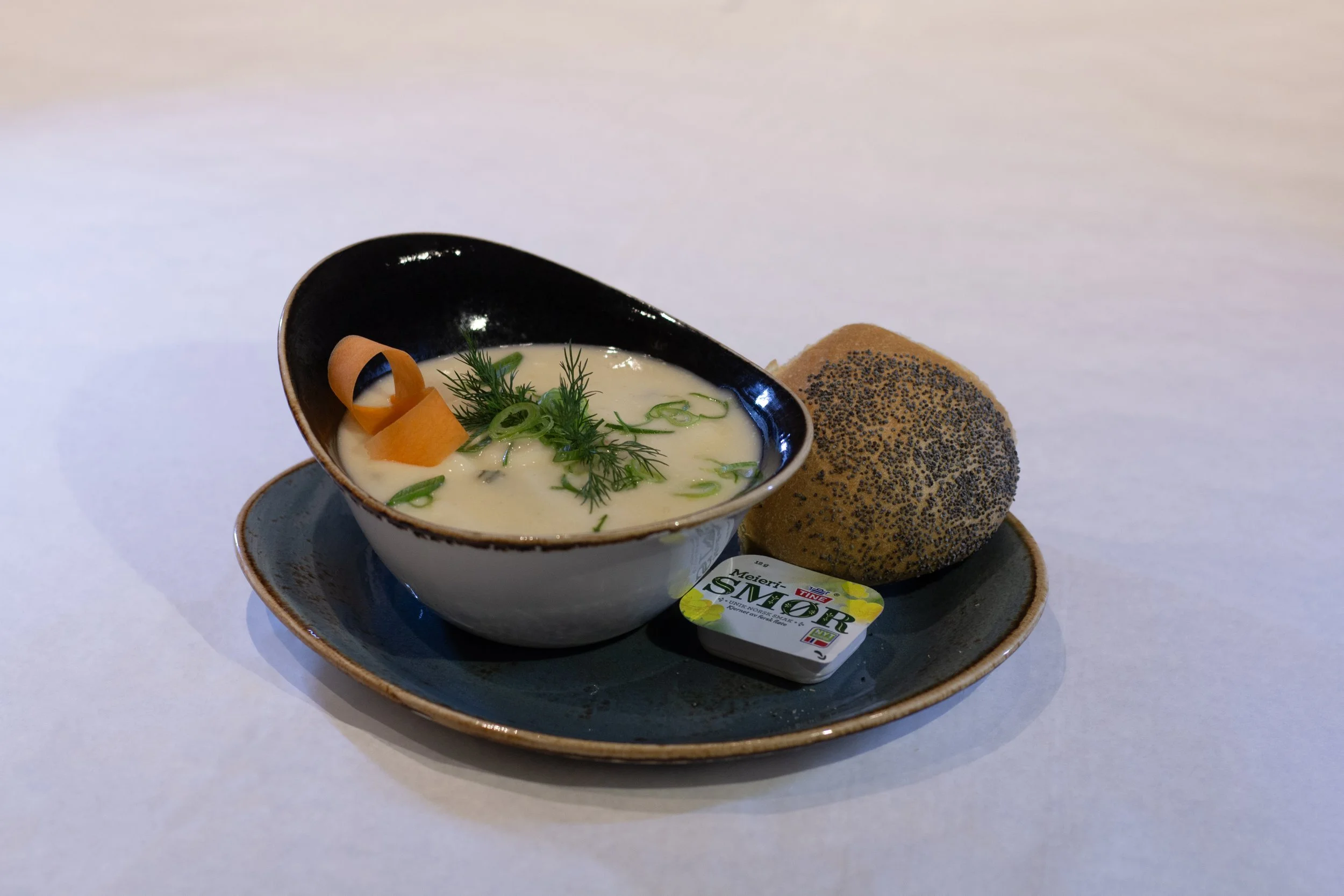 A bowl of creamy soup garnished with fresh herbs and sliced green onions, accompanied by a seeded bread roll, on a dark plate with a small packet of margarine