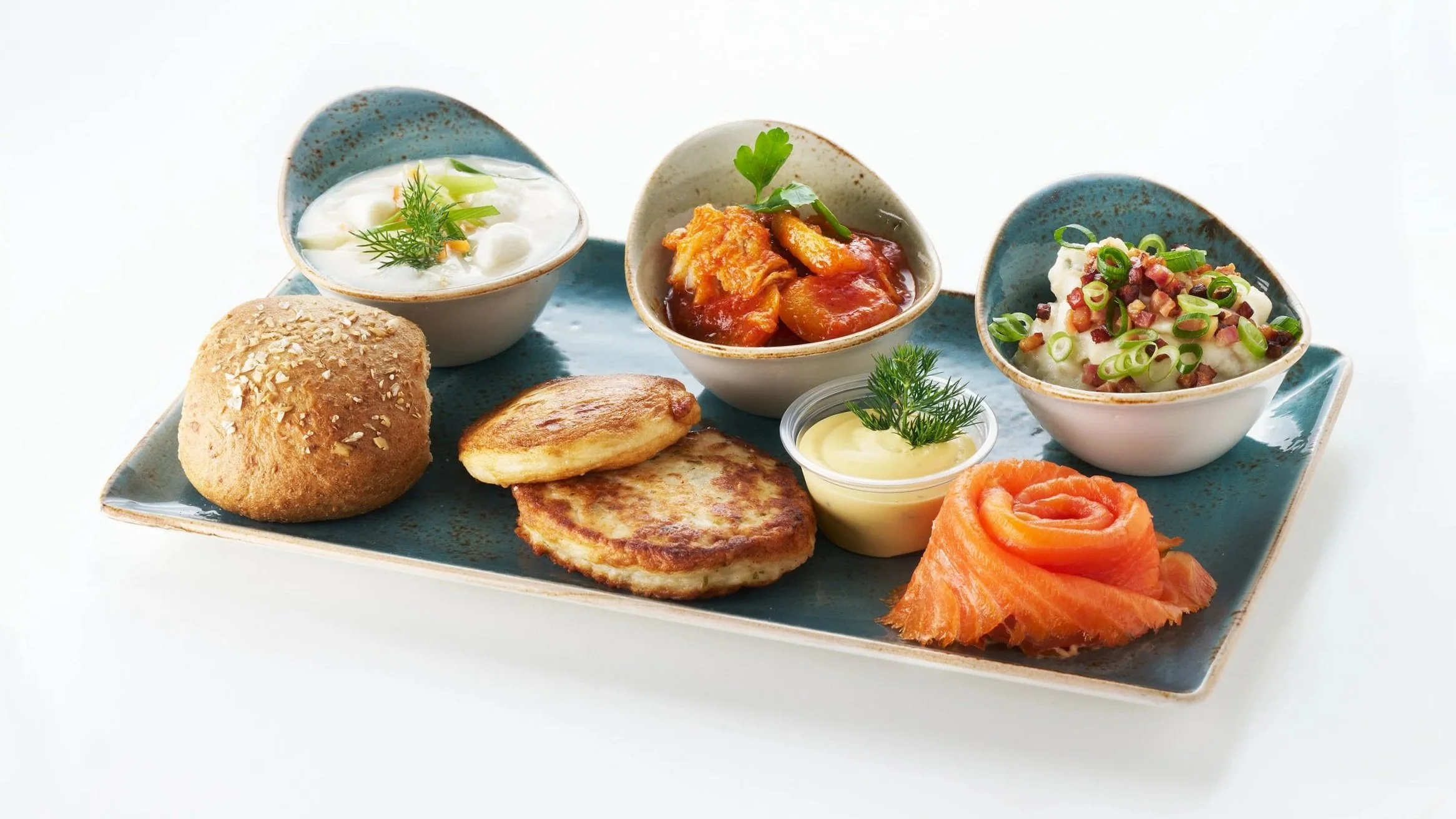 A plate of assorted breakfast foods including bread roll, hash brown, smoked salmon, a small appetizer with smoked salmon on top, and three bowls of different toppings or side dishes, such as sour cream with herbs, stew with vegetables and meat, and a potato salad.
