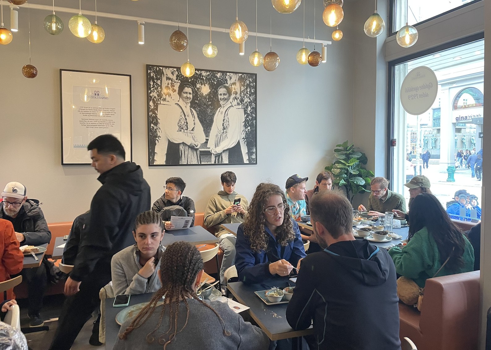 People dining at a busy restaurant with large black and white photos on the wall and a large window showing an outside city street.
