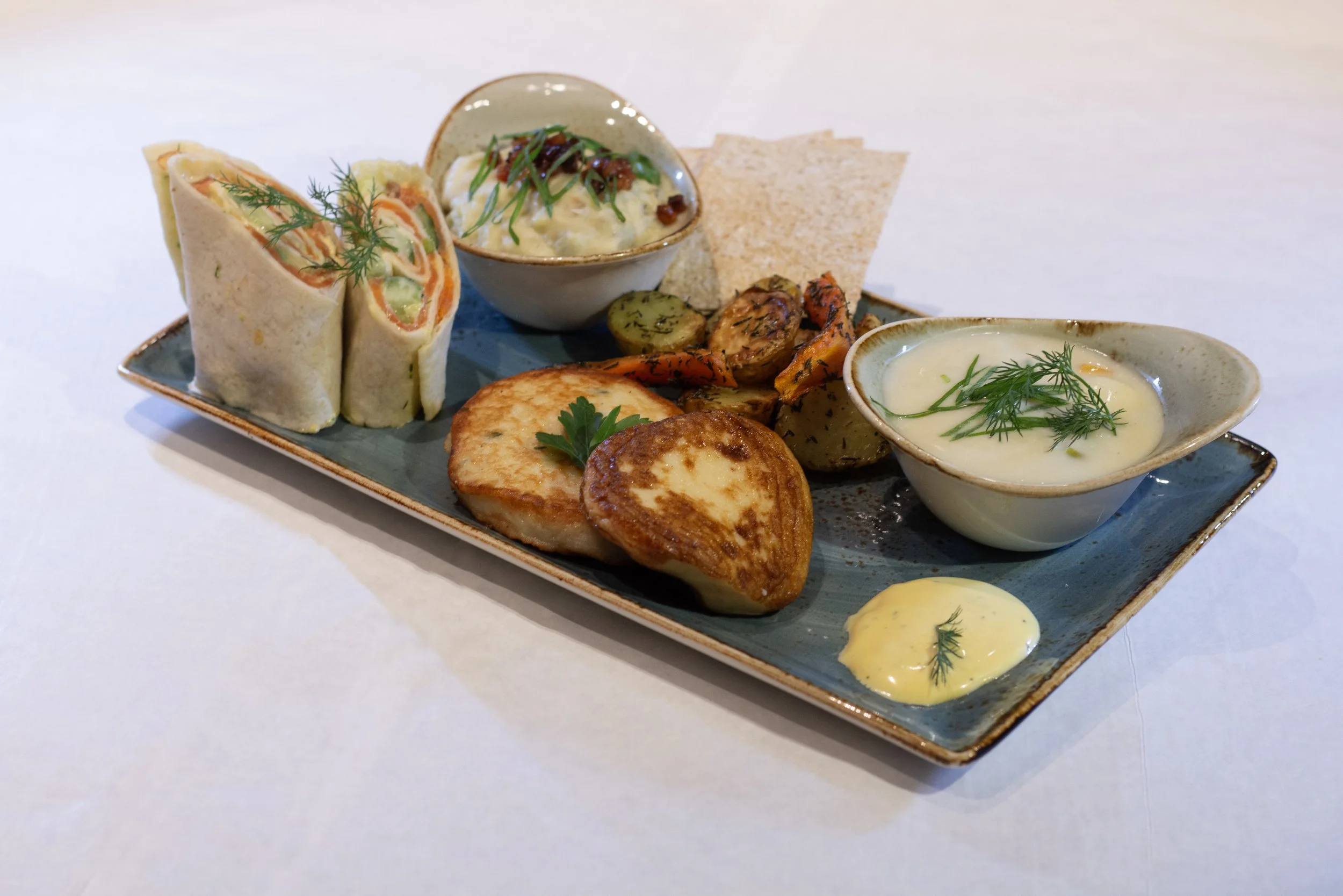 A rectangular ceramic plate with Indian food including wraps, roasted vegetables, shallow bowls of creamy sauces garnished with herbs, and a few chips.