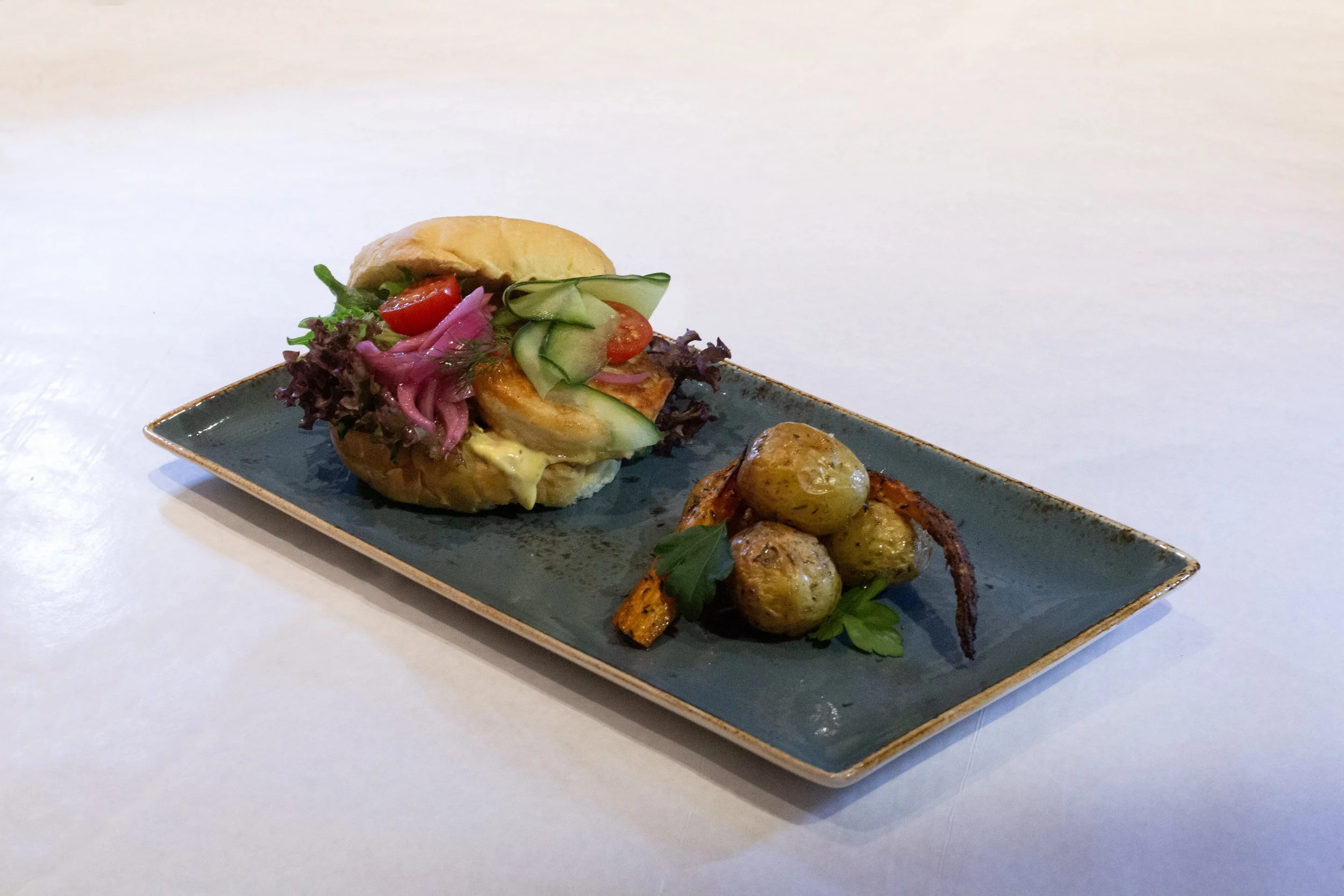 A rectangular plate with three types of food: a small sandwich with greens and vegetables inside, roasted vegetables with a skewer, and two potatoes topped with sauce.