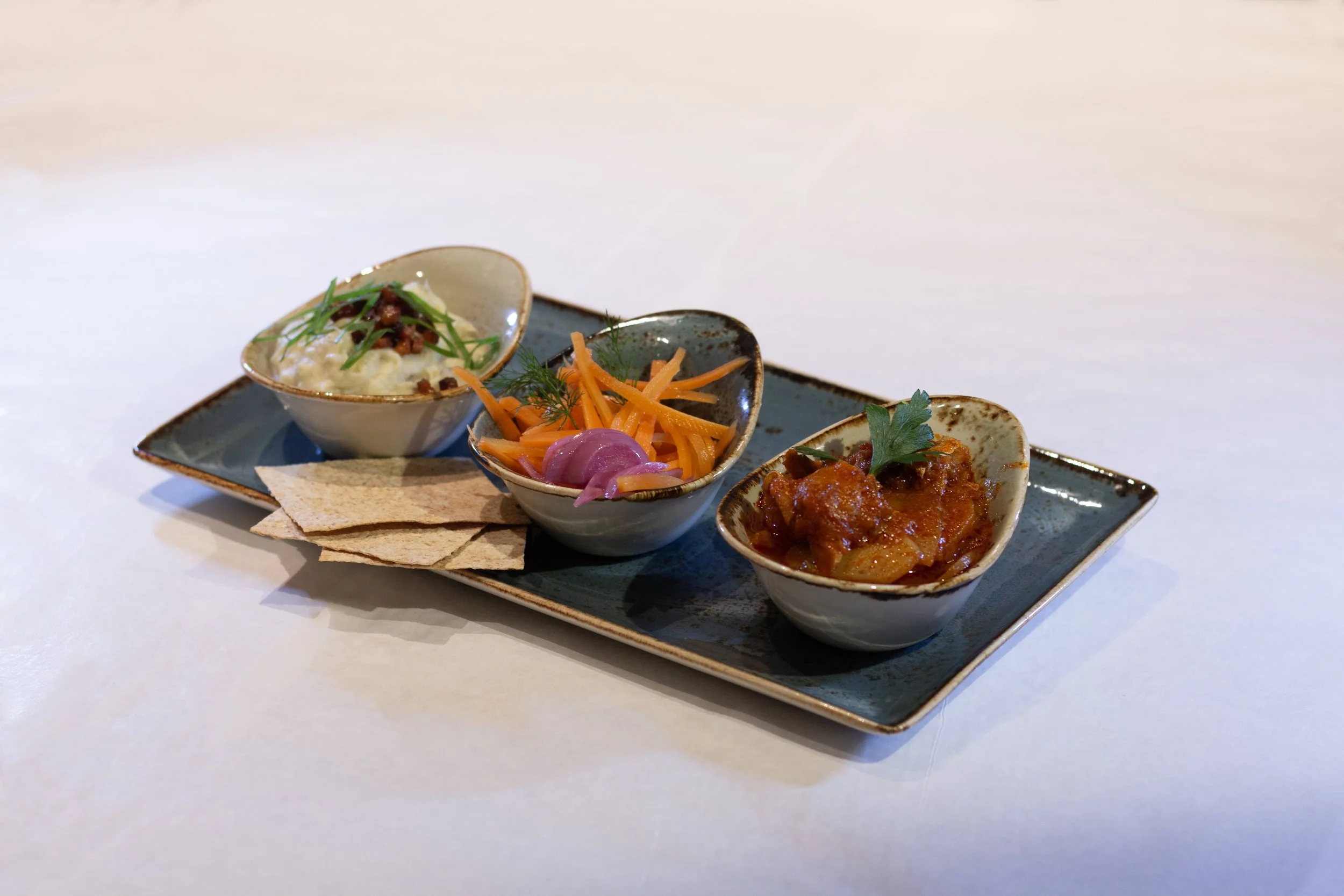 A rectangular ceramic plate holds three small bowls of different Indian dishes with crackers on the side, on a white surface.