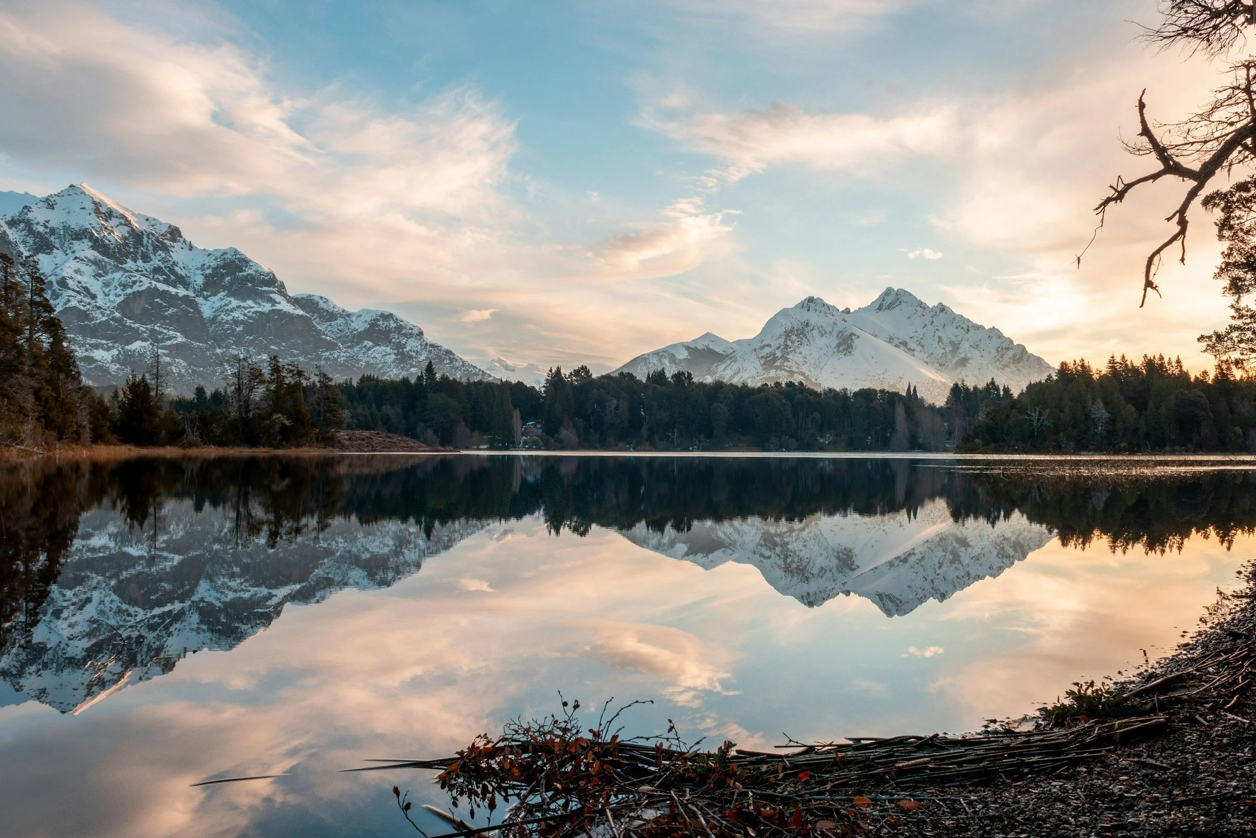 A tranquil lake reflecting snow-covered mountains and a partly cloudy sky at sunset or sunrise.